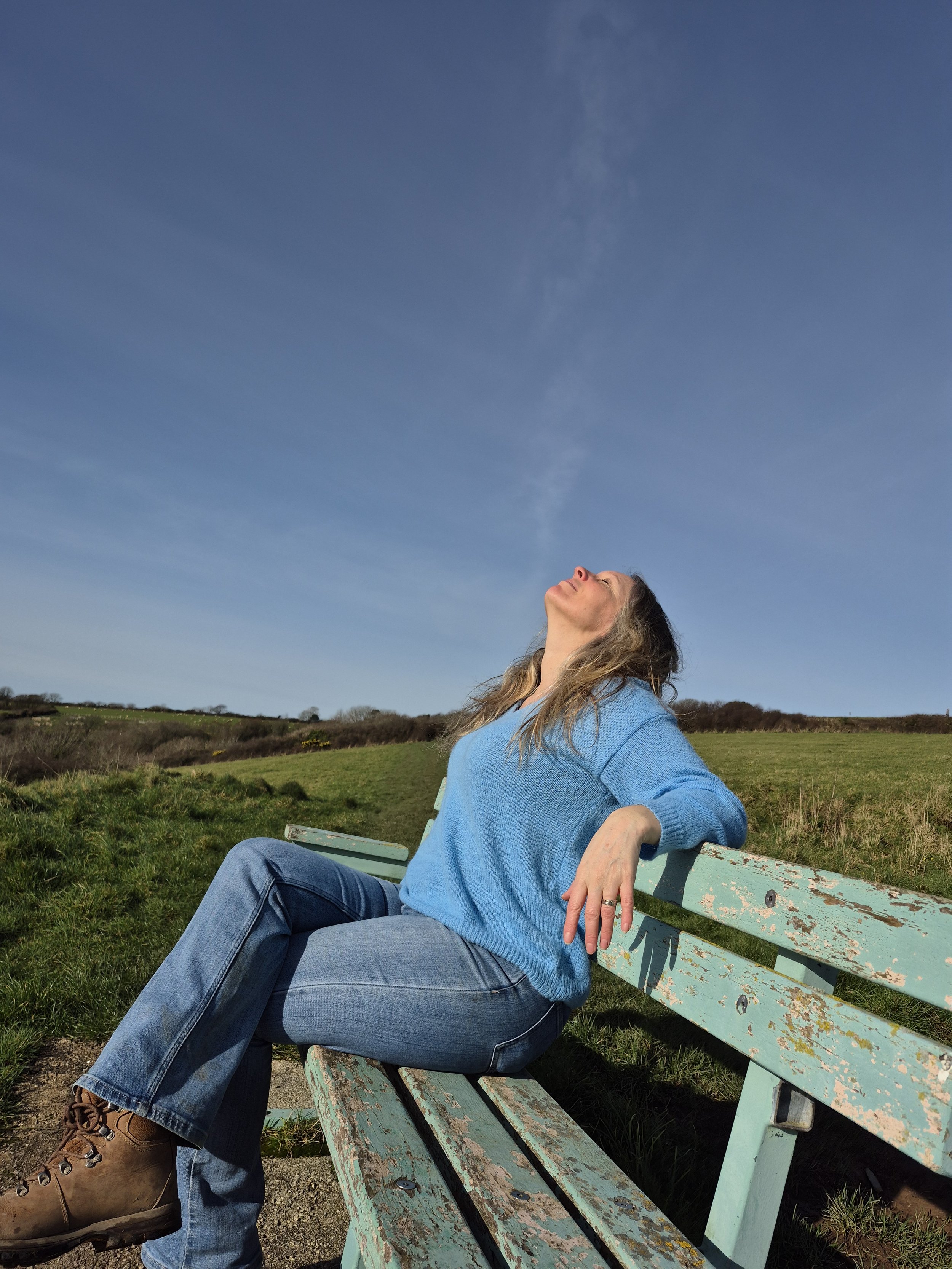 Laura Jessica walker sitting on a weathered green park bench outdoors, enjoying sunlight with her eyes closed and face tilted upward, against a blue sky and open grassy landscape.