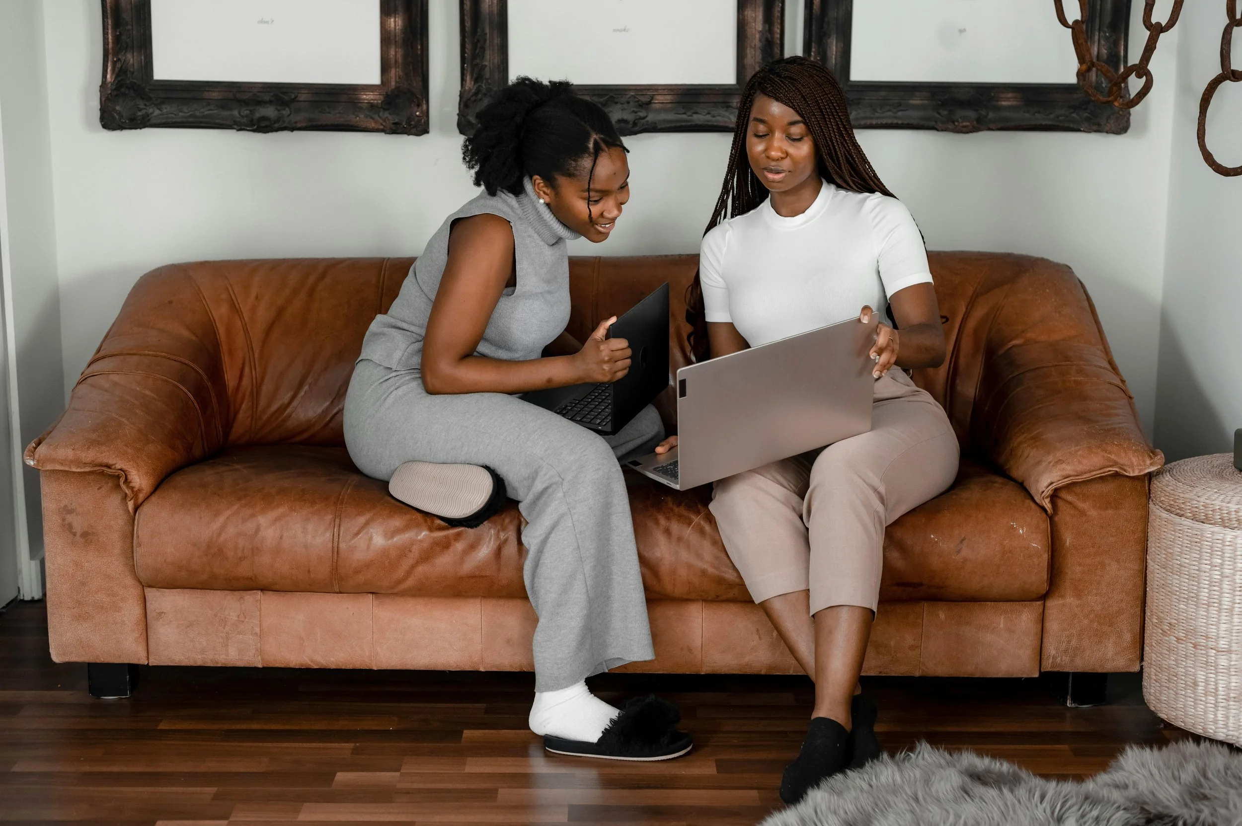 Two young women sitting on a couch with their laptops and chatting