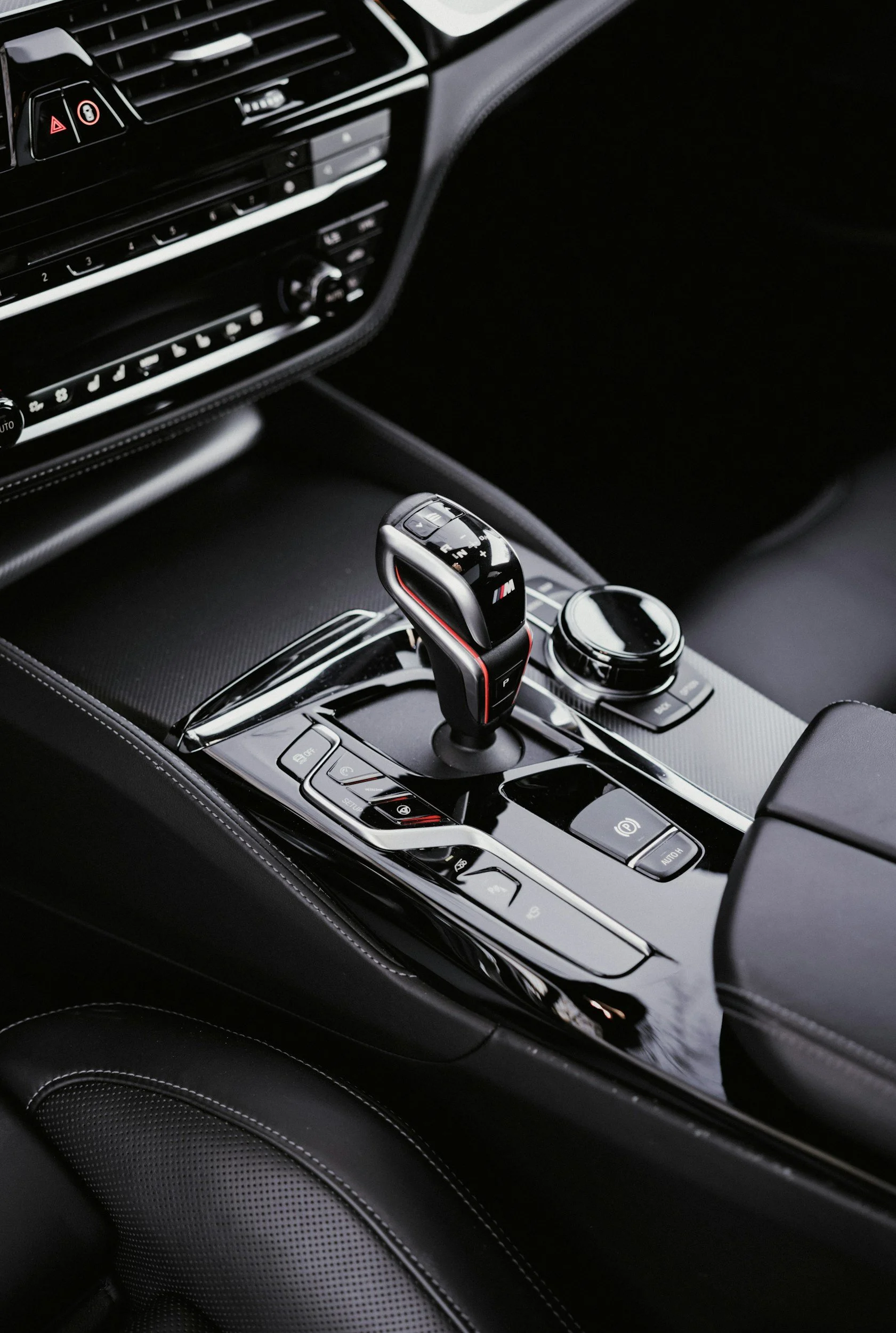 Close-up of a BMW car gear shift lever, surrounded by a sleek, black center console with various control buttons and a circular dial. The interior features black leather with white stitching.