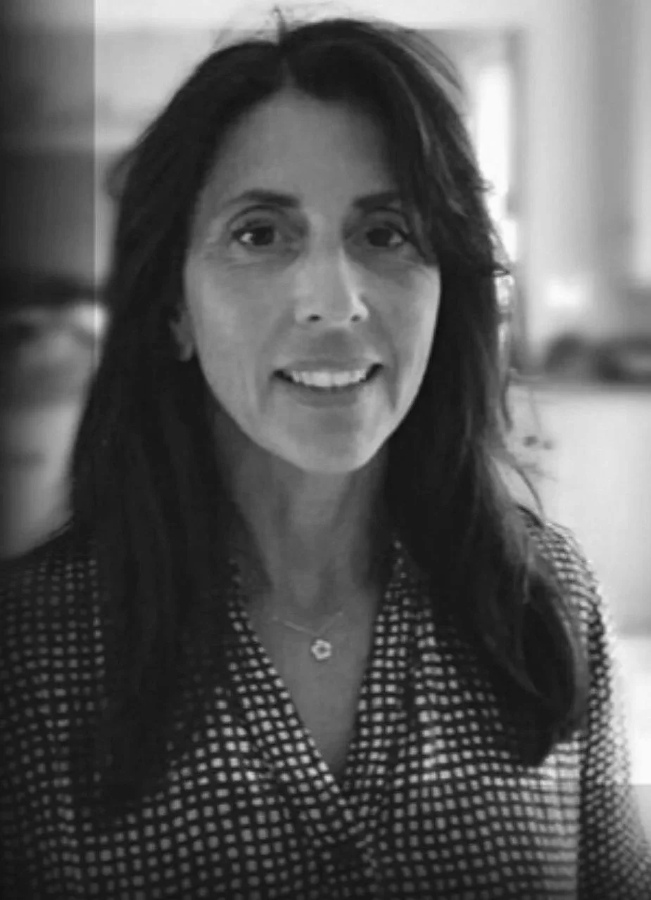 A woman with long dark hair smiling, wearing a checkered shirt and a necklace, in an indoor setting.