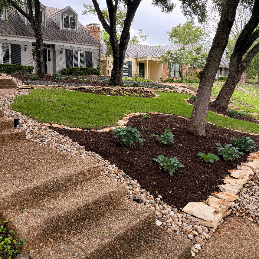 Residential front yard with stone steps, green grass, trees, and landscaped beds with shrubs and mulch, in a suburban neighborhood.