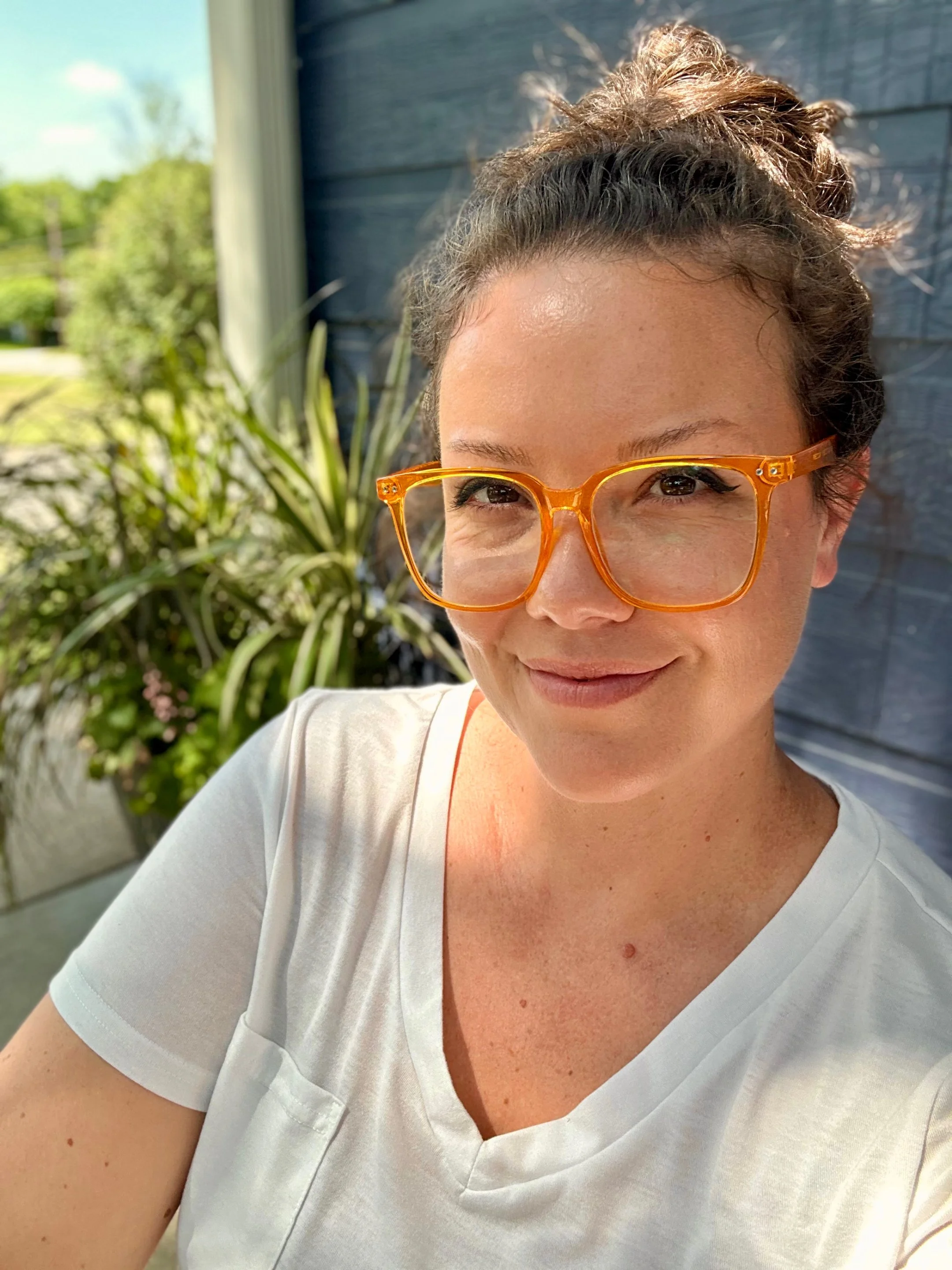 A woman with curly hair in a bun wearing orange glasses and a white shirt, smiling outdoors in sunlight with greenery and a blue wall in the background.