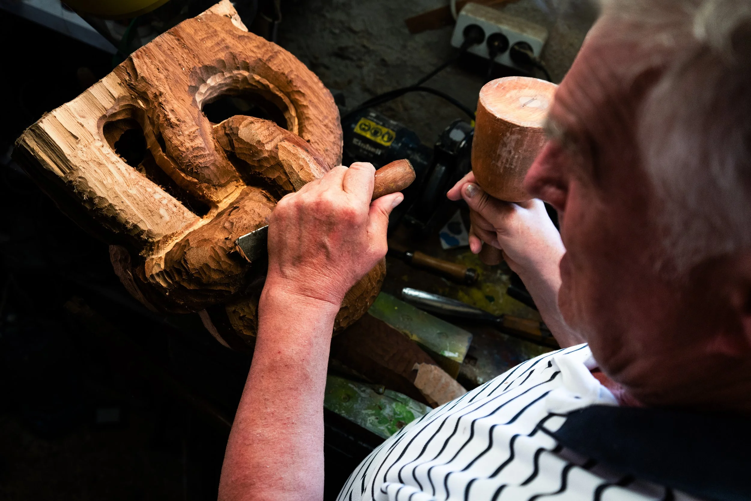 A person carving a wooden mask during a woodworking project, with various tools and a power outlet in the background.