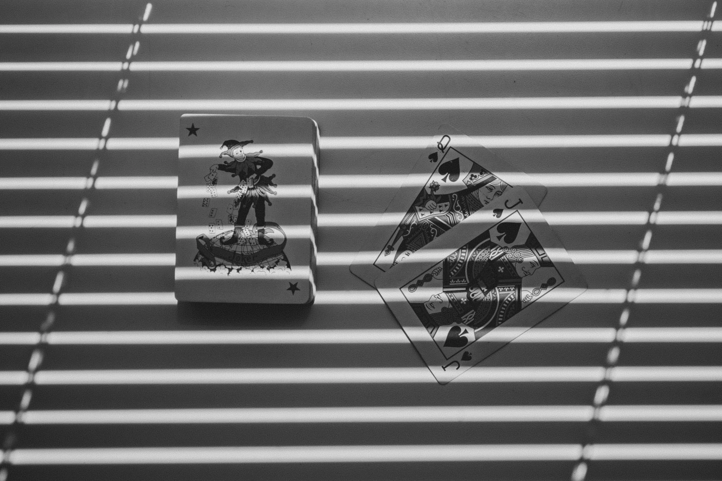 A deck of Joker playing cards and a spread of four Queen of Spades playing cards on a surface with horizontal blinds casting shadows.