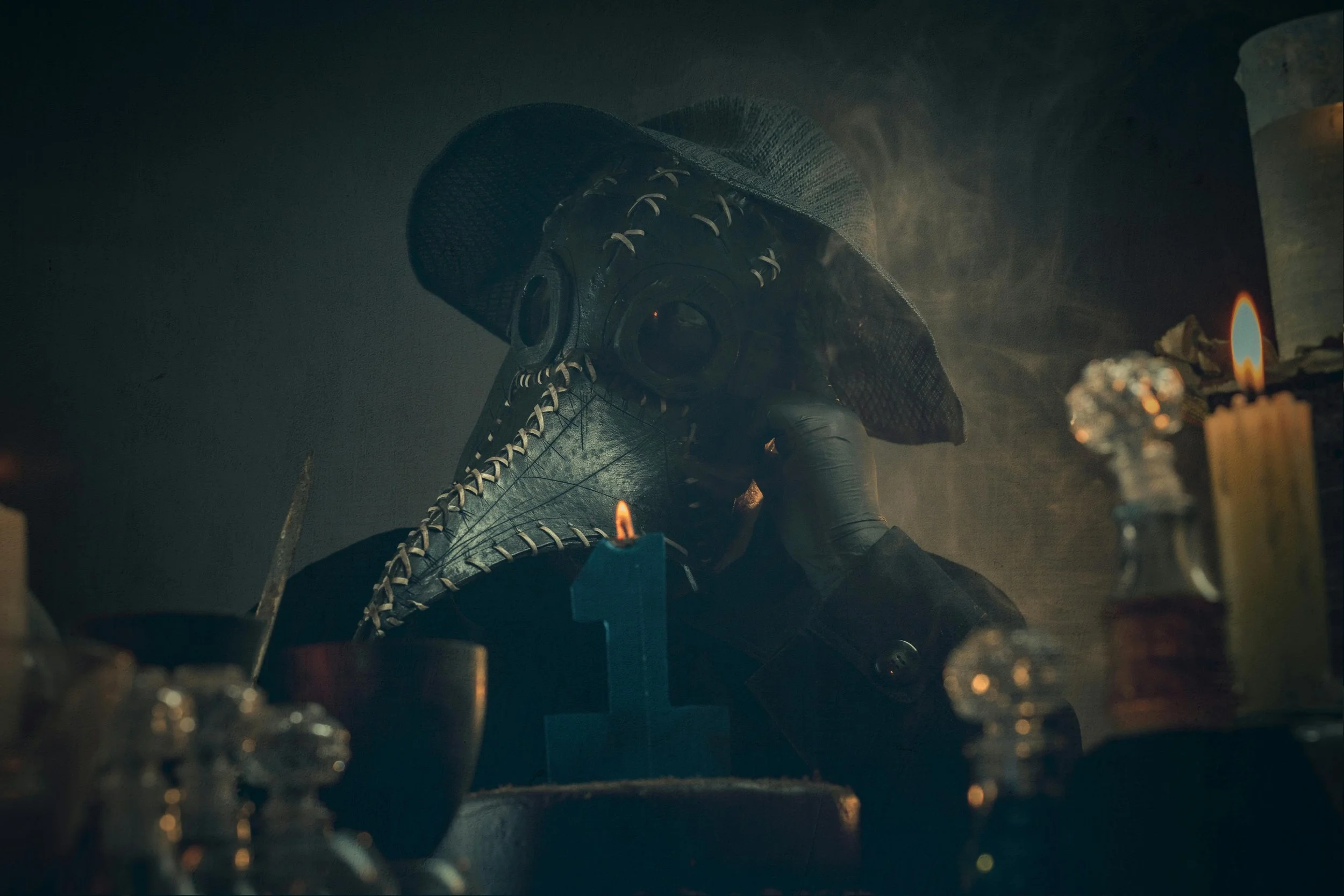 Person wearing a plague doctor mask with a tall bird beak, sitting at a table with candles and bottles, in a dimly lit room.