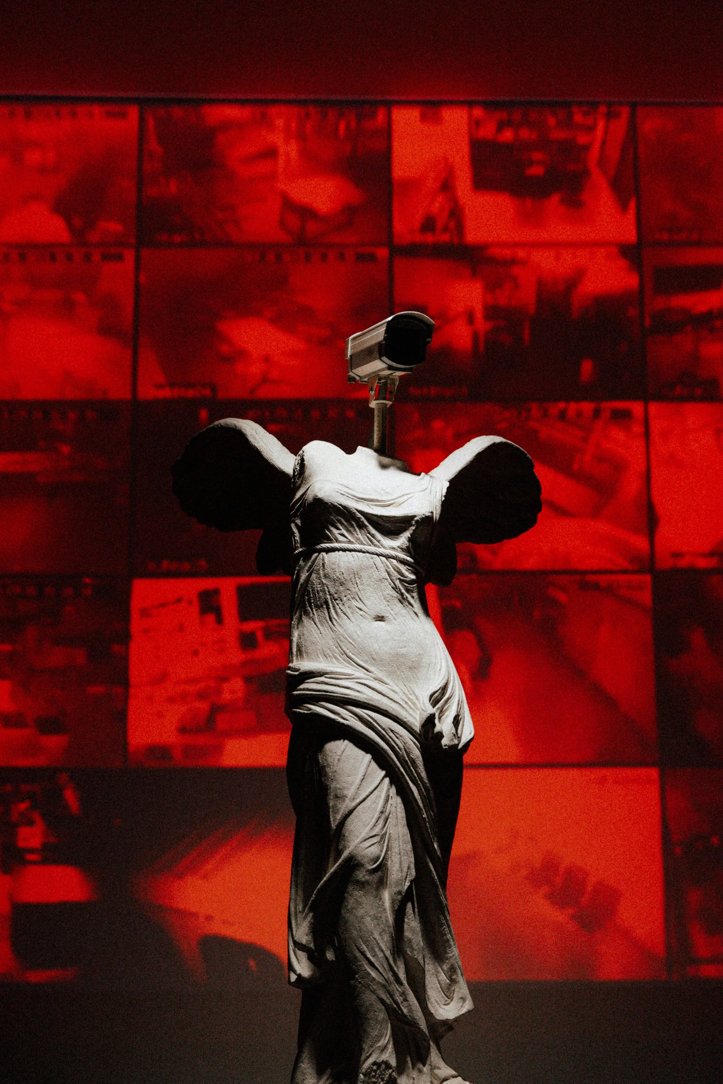A classical statue of a woman with broken head, with a security camera mounted on her back, in front of a red background with multiple images displayed on screens.