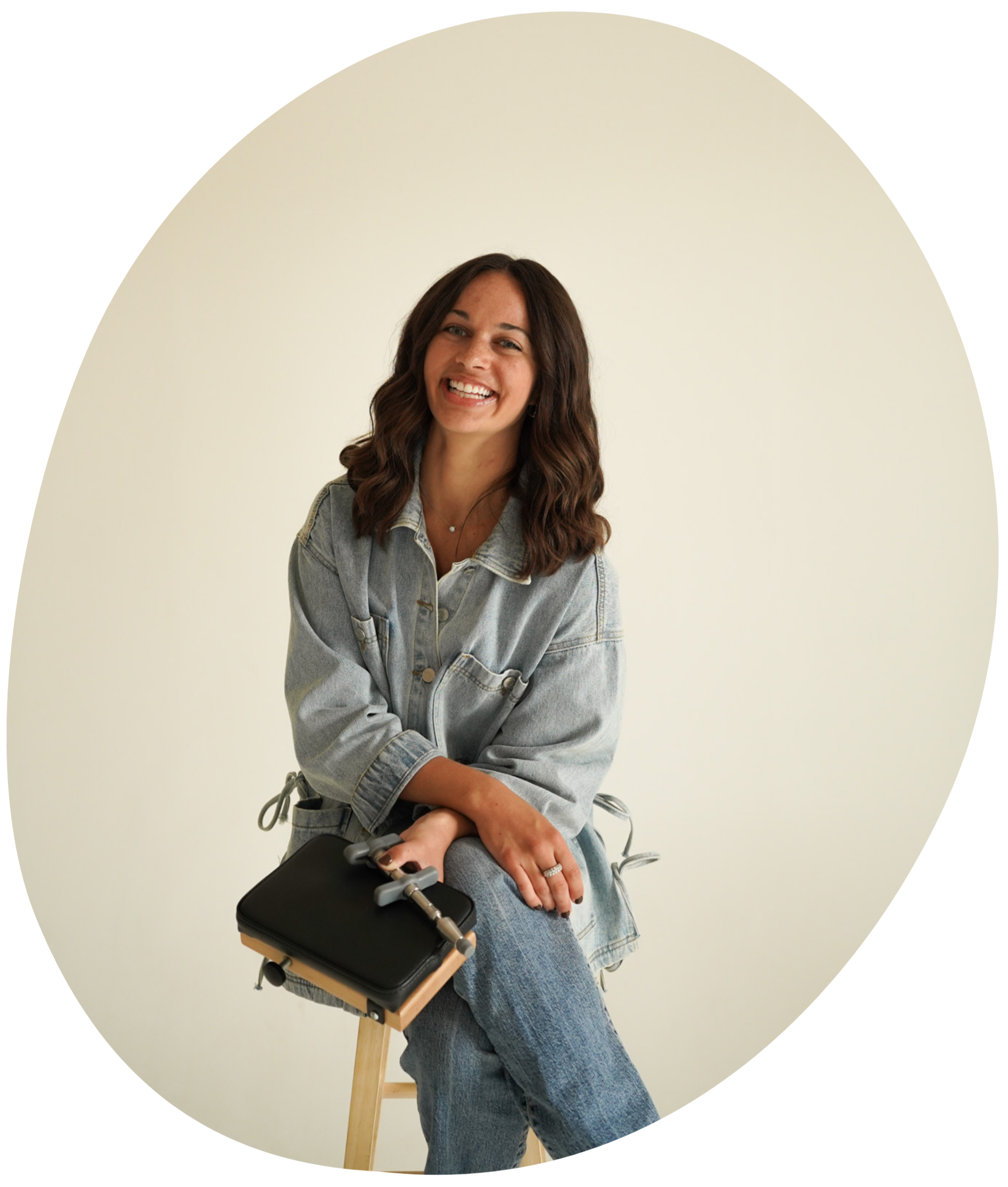 A woman with brown wavy hair smiling, sitting on a wooden stool, wearing a light denim jacket and jeans, holding a black notebook and pen, against a plain light background.
