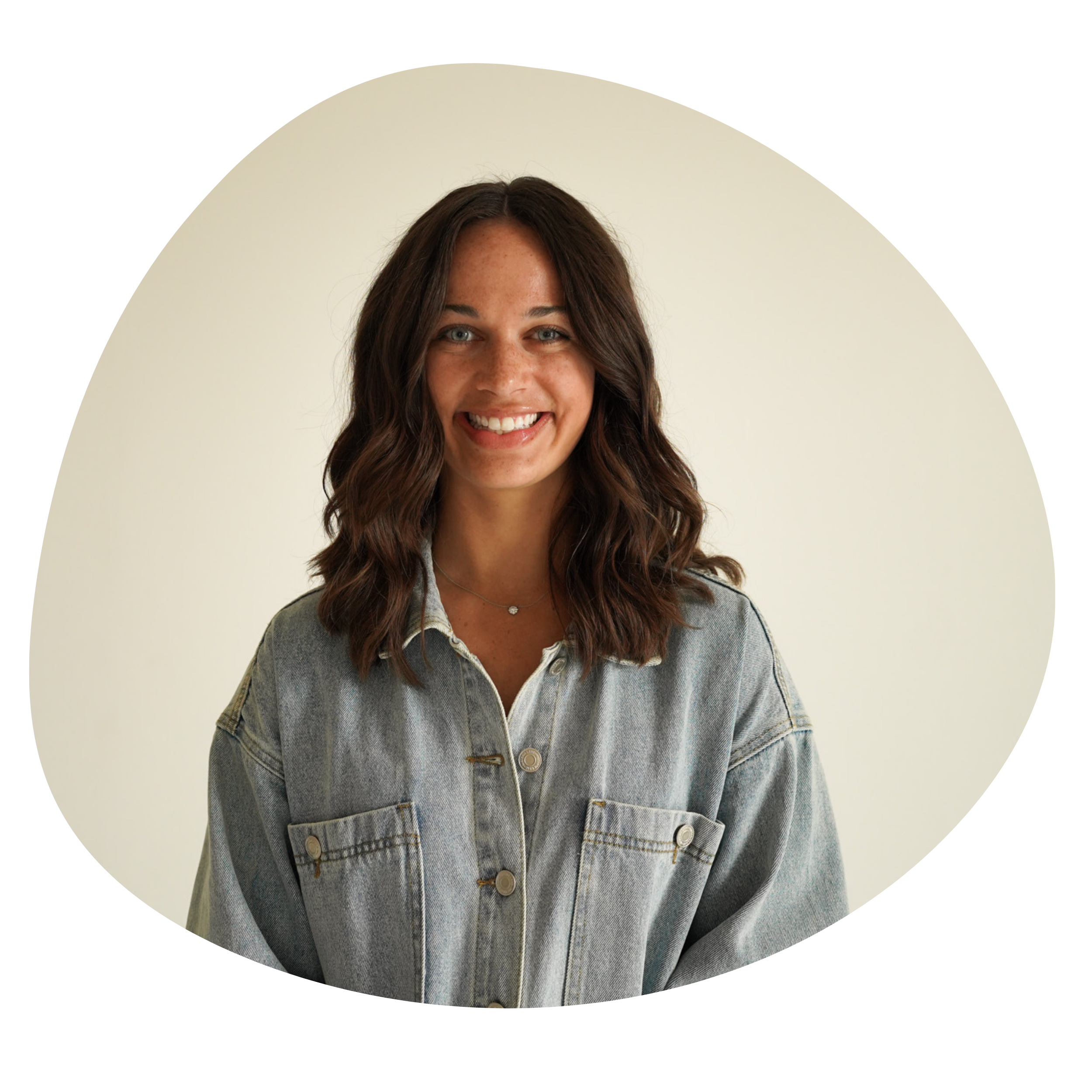 A young woman with wavy brown hair, blue eyes, and freckles, smiling and wearing a light blue denim jacket over a white shirt, standing against a plain beige background.