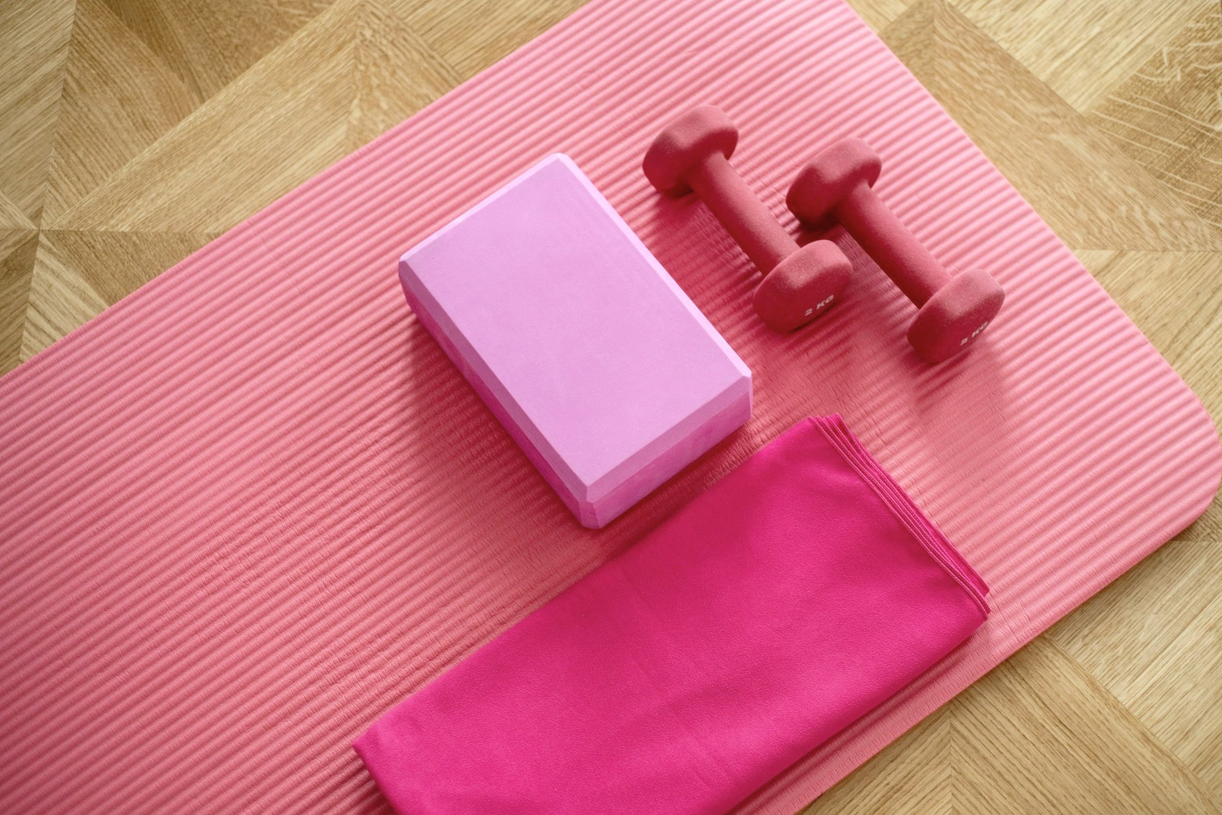 A pink yoga mat with a pink foam block, pink small dumbbells, and a pink resistance band on a wooden floor.
