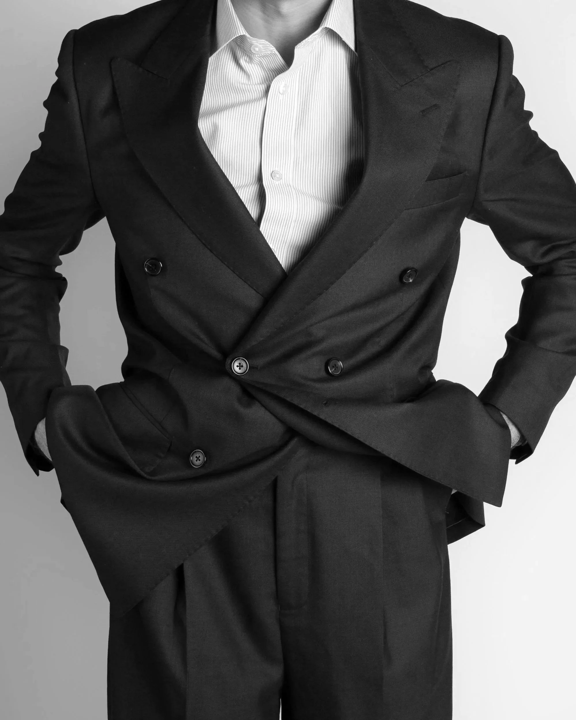 A person wearing a black double-breasted suit jacket and a white collared shirt, with their hands on their hips, in front of a plain background.