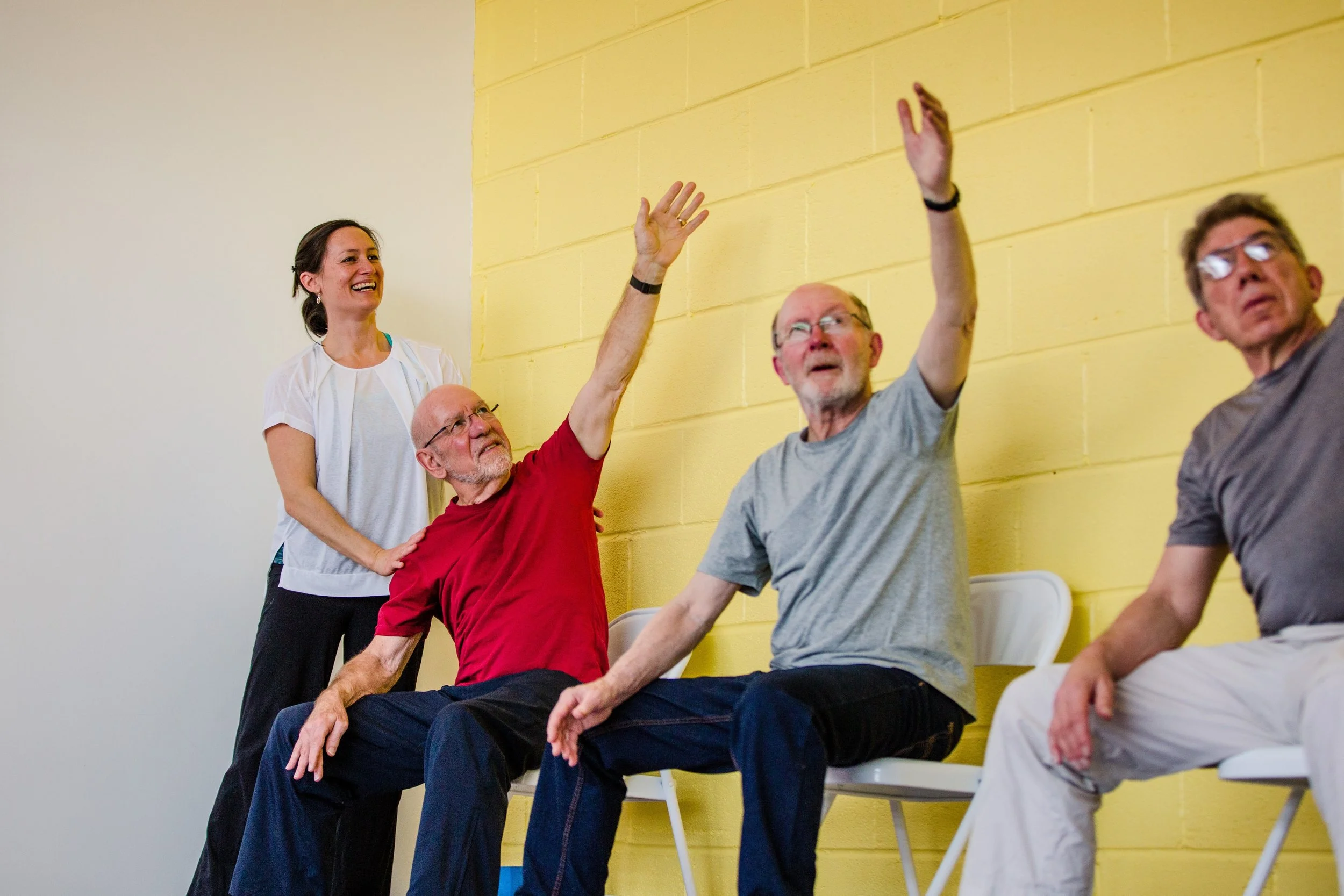 Group of elderly people in a seated exercise class, with a woman standing and a man raising his hand, all smiling and engaging in activity against a yellow wall.