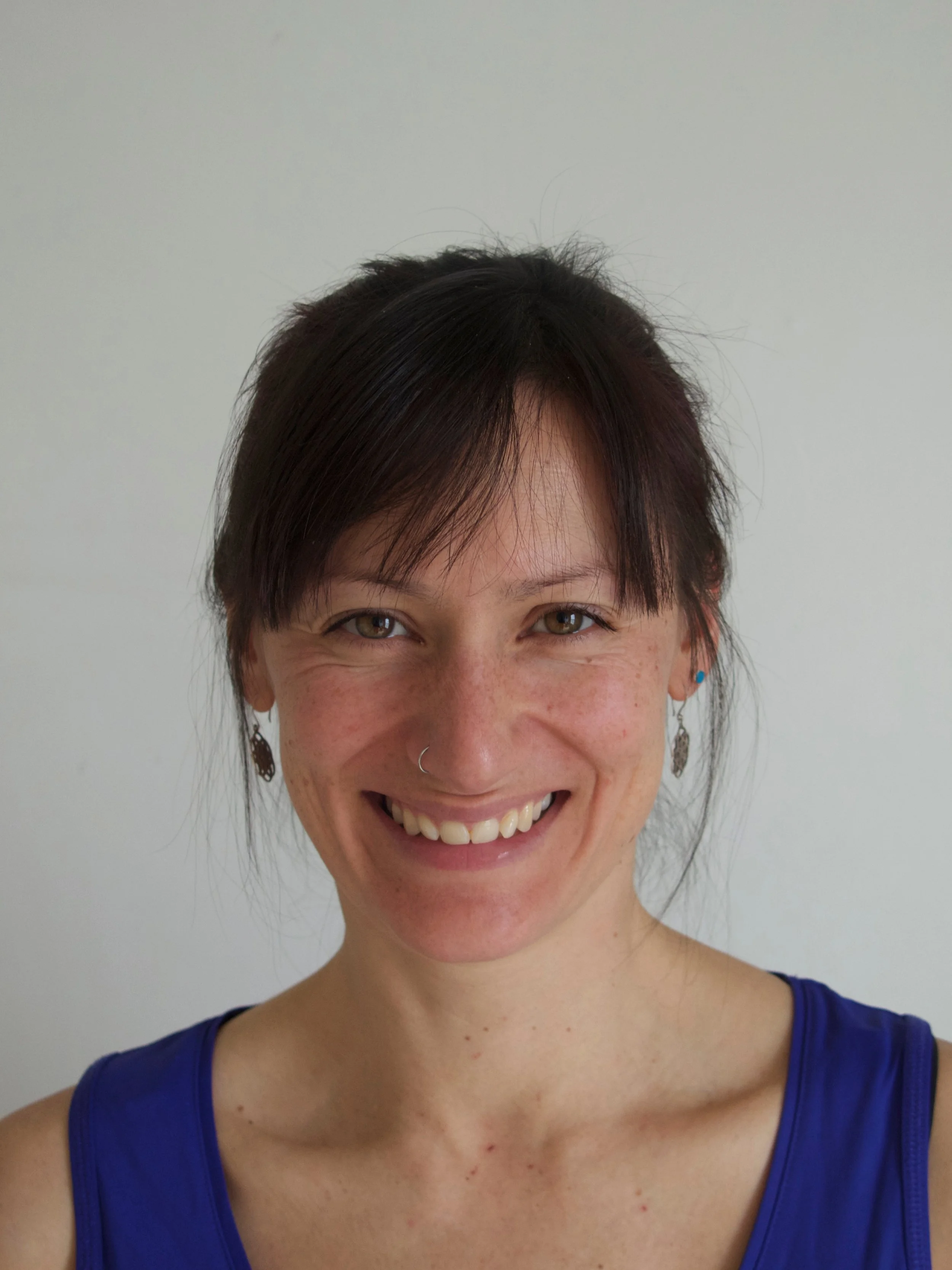 A woman with short dark hair, wearing a blue tank top, smiling, with earrings and a nose ring, standing against a plain background.