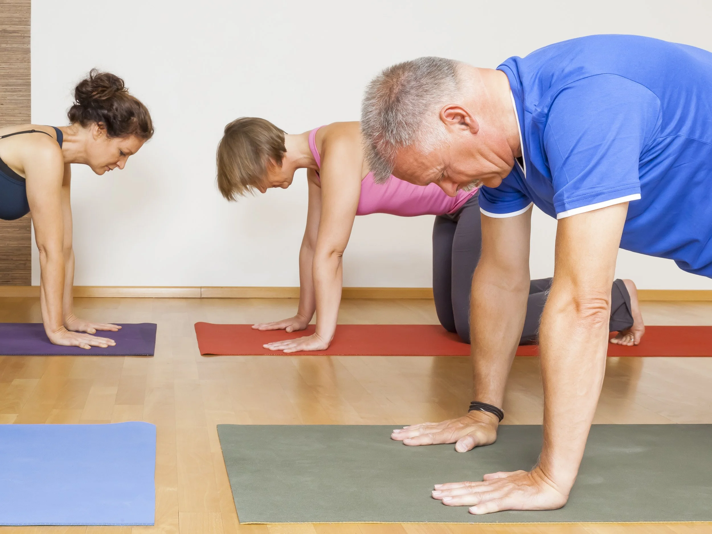 Three people practicing yoga in downward dog pose on colorful mats in a studio.