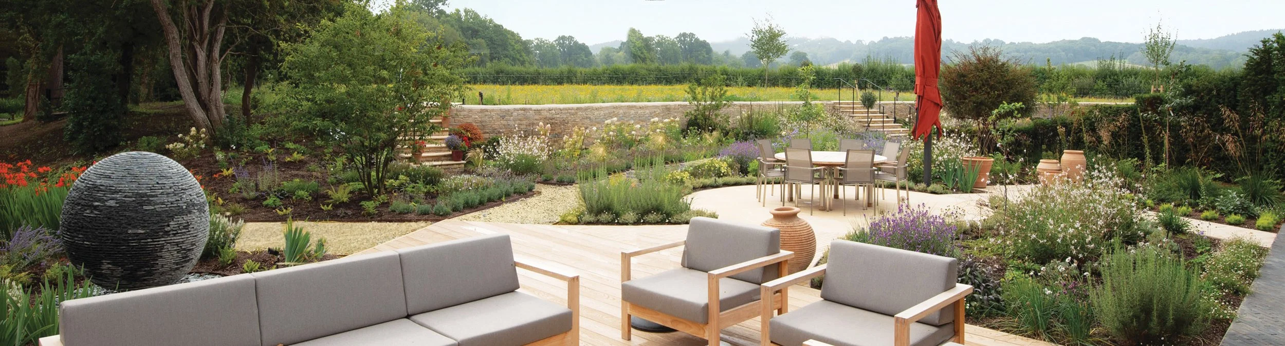 Landscape garden designed by Doug Holloway, showing the main seating area, water feature, Millboard decking, London Stone paving and spiralling naturalistic planting, with views towards the surrounding countryside.