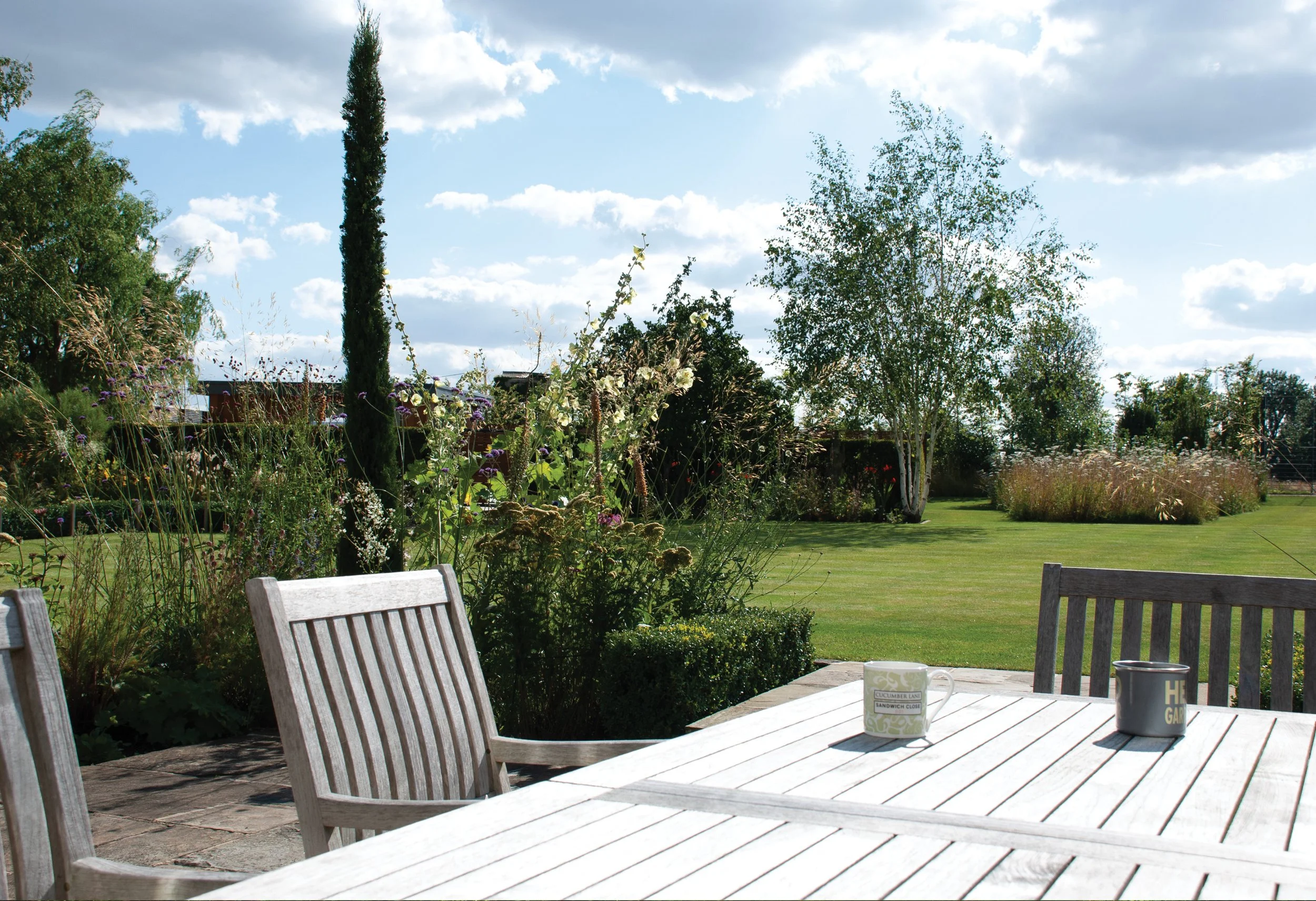 Relaxed dining terrace enclosed by planted beds, with Stipa gigantea, upright junipers and views towards birch trees and wildflower planting.