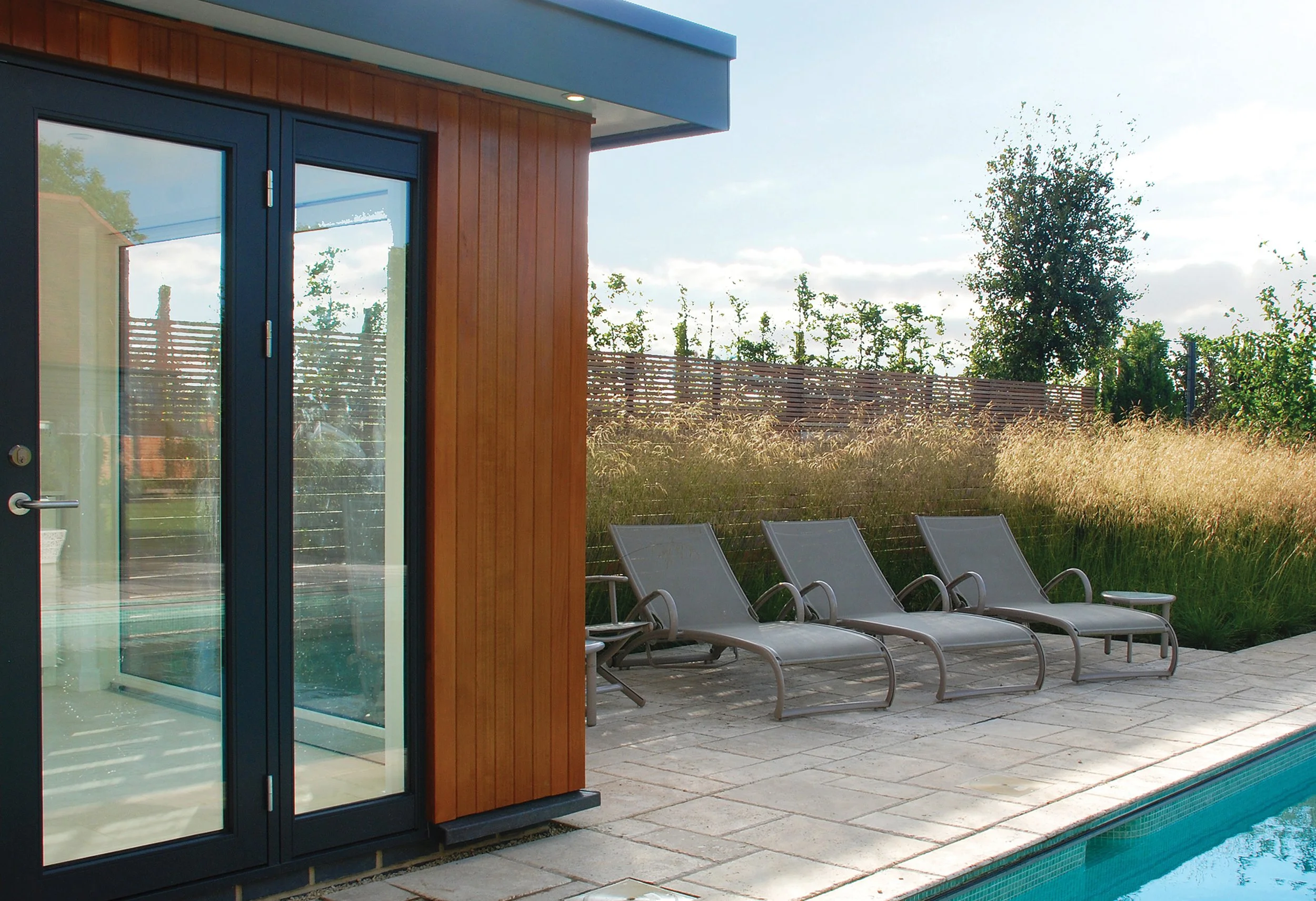 Cedar pool house with aluminium doors and windows, with restrained grass planting creating a simple contemporary poolside space.