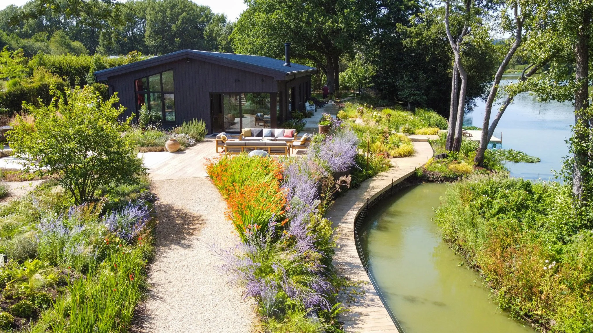The contemporary black timber house and curving boardwalk by stream, surrounded by colourful naturalistic planting beside the lake.
