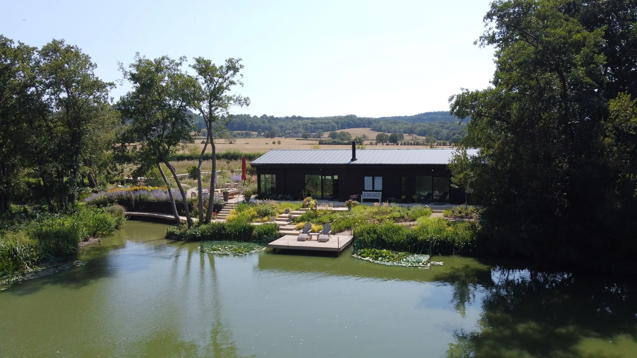 View across the lake towards The Lake House, with floating 'Lillypad' decking steps, waterside planting and mature trees.