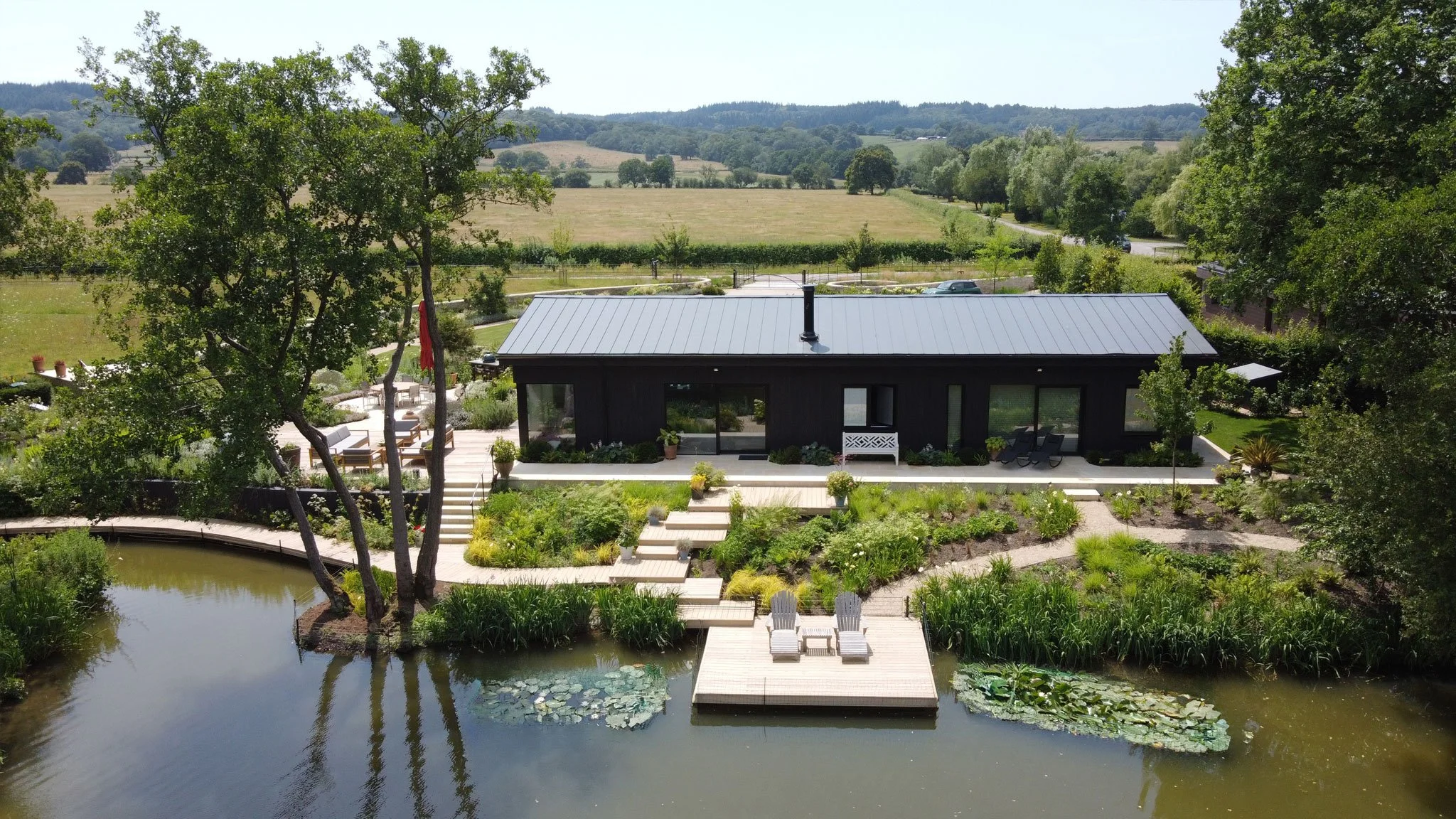 The Lake House seen across the water, with naturalistic planting, floating deck and mature trees framing the contemporary house.