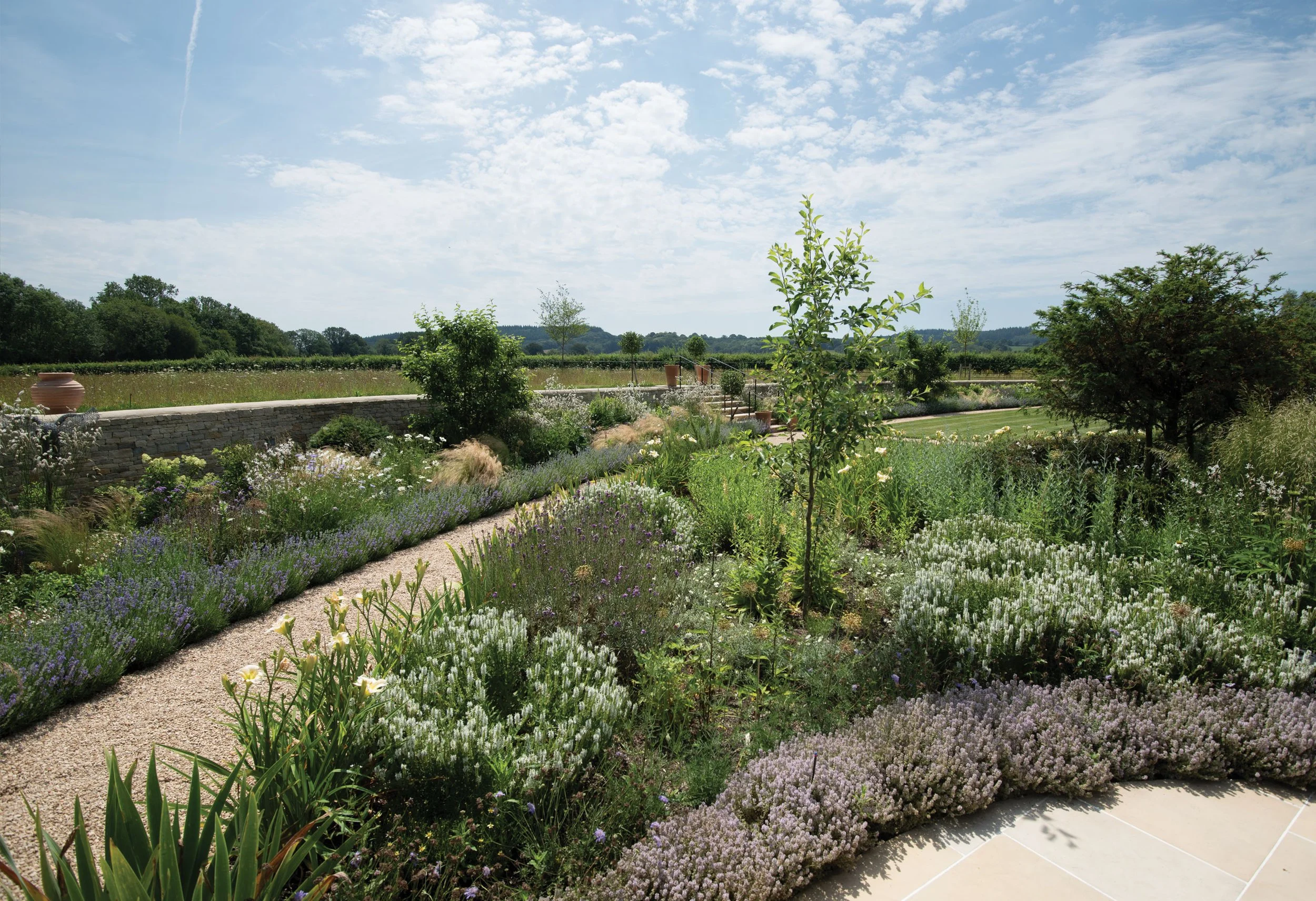 Naturalistic perennial planting beside a curving gravel path, with meadow and open countryside beyond.