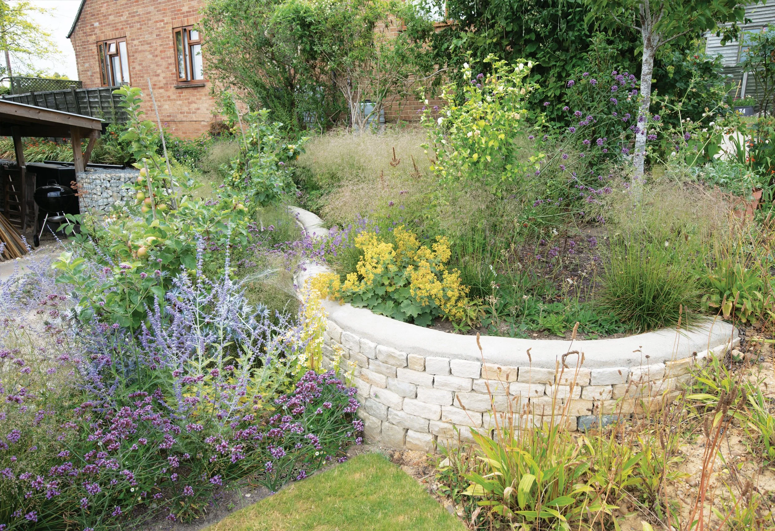 Curving dry-stone wall enclosing abundant naturalistic planting beside the lawn and terrace.