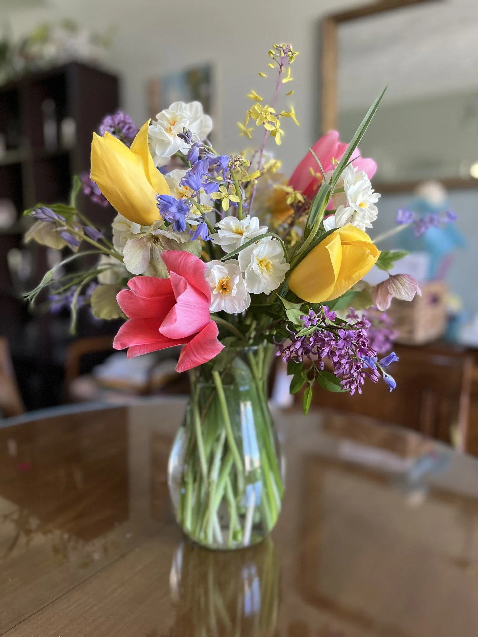 Colorful bouquet of yellow, pink, white, purple, and blue flowers in a clear glass vase on a wooden table.