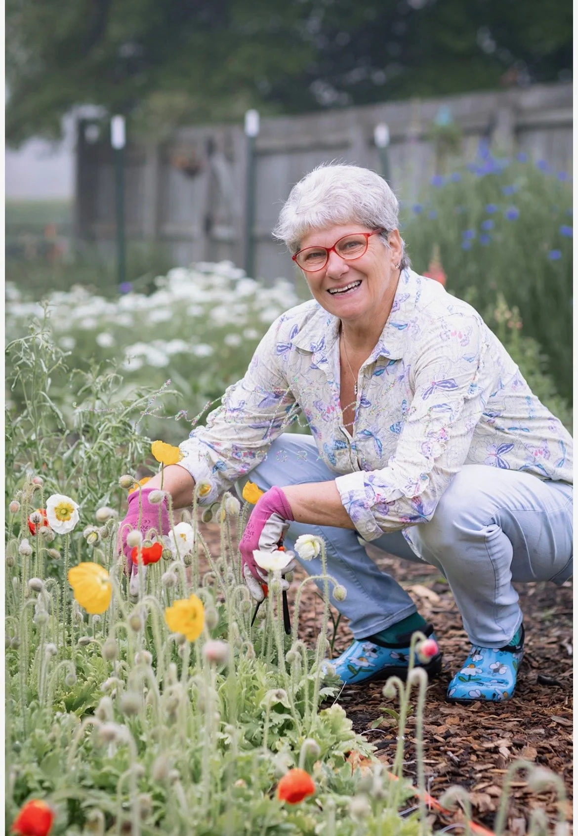 An elderly woman with gray hair, red glasses, and wearing gardening gloves is gardening in her yard, smiling while tending to colorful flowers.