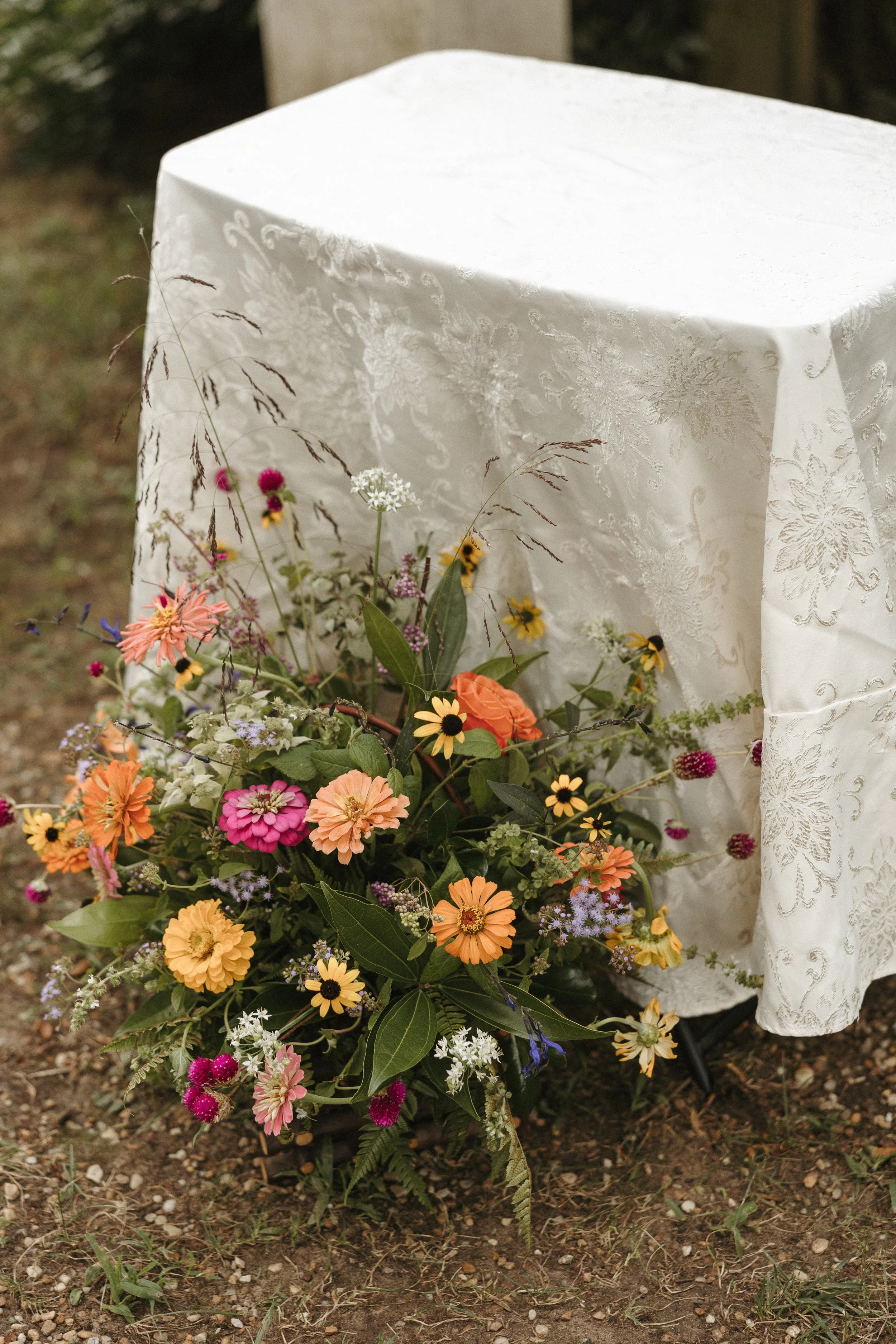 Flower arrangement with various colorful flowers placed on the ground in front of a white embroidered tablecloth covering a table.