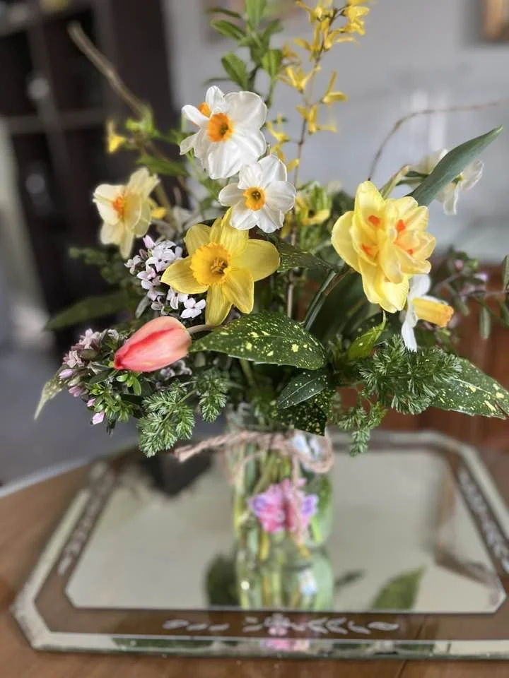 A bouquet of colorful flowers in a clear glass vase placed on a reflective tray on a wooden surface.