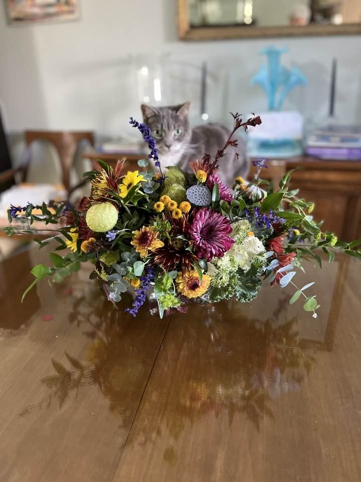 A colorful floral arrangement on a wooden table with a cat sitting behind it.