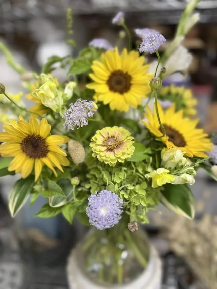 A colorful bouquet of yellow sunflowers, purple scabiosa, and other assorted flowers in a glass vase.