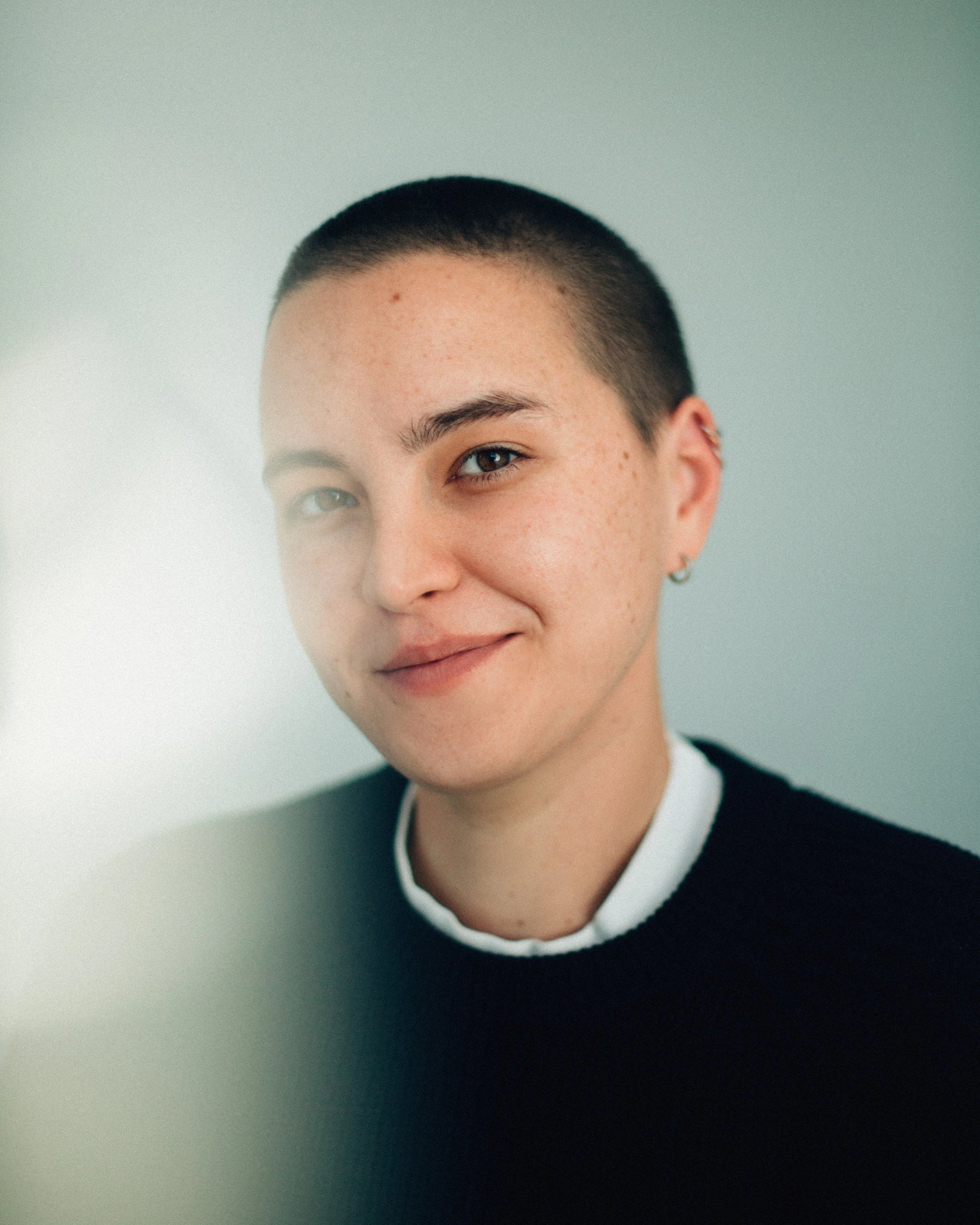 Close-up of a smiling young woman with short, dark hair, wearing a black sweater and a white shirt collar, standing against a plain, muted background.