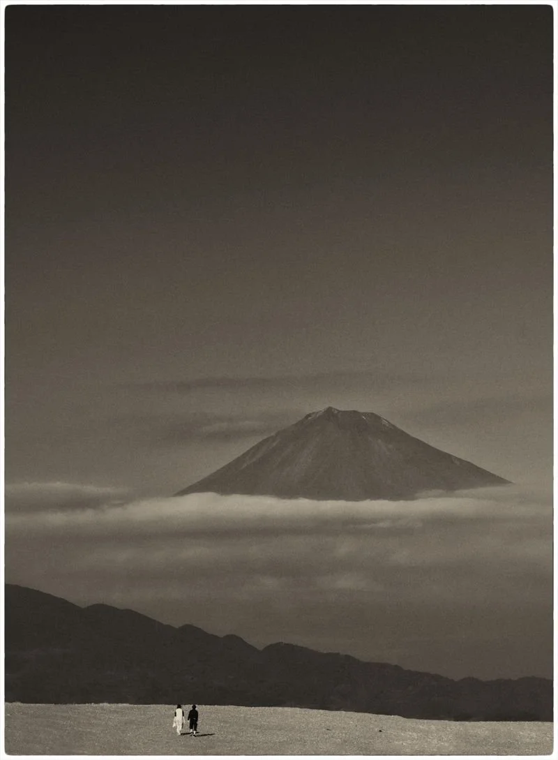 A black and white photo of Mount Fuji with clouds surrounding it, and two small people walking in the foreground.