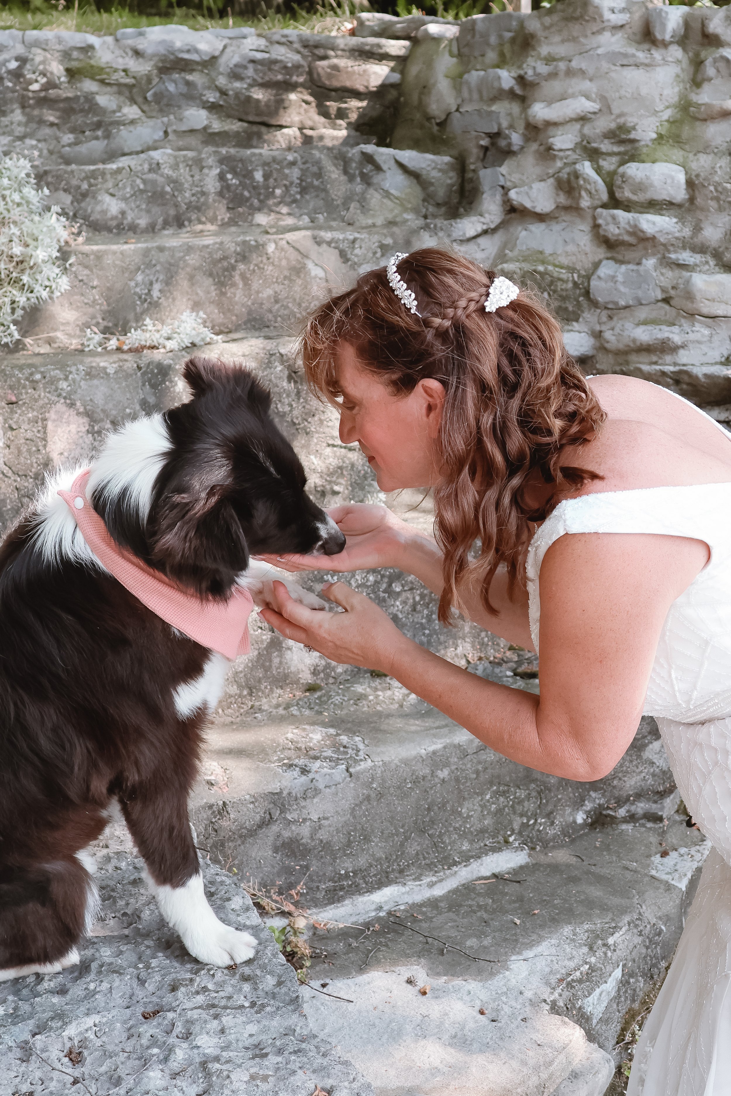 A woman with curly hair, wearing a white dress and a beaded hair accessory, is bending down and gently holding the face of a black and white Border Collie dog. They are outdoors on a stone staircase with a stone wall in the background.