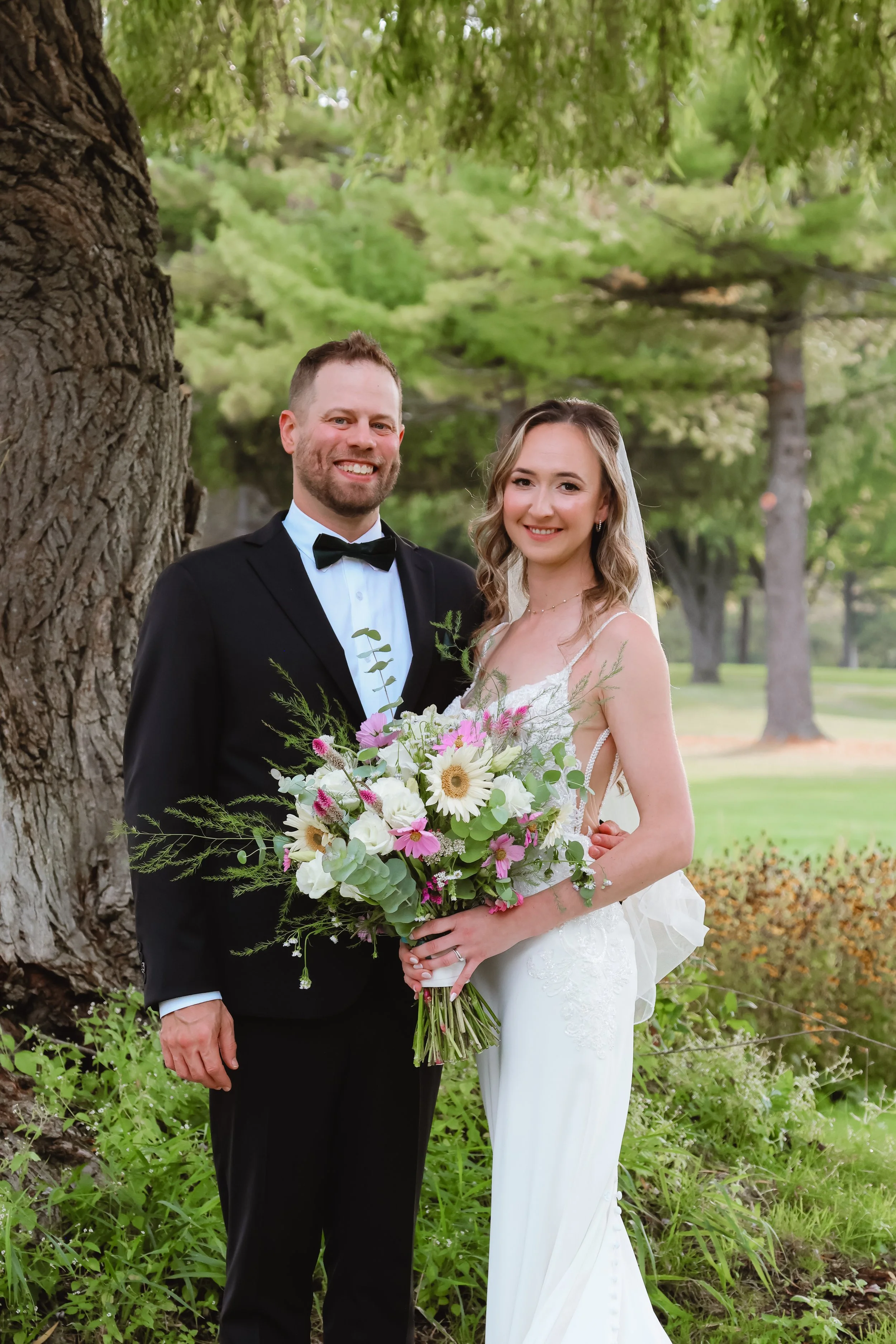 A smiling bride and groom standing outdoors by a large tree, holding a floral bouquet, with greenery and other trees in the background.