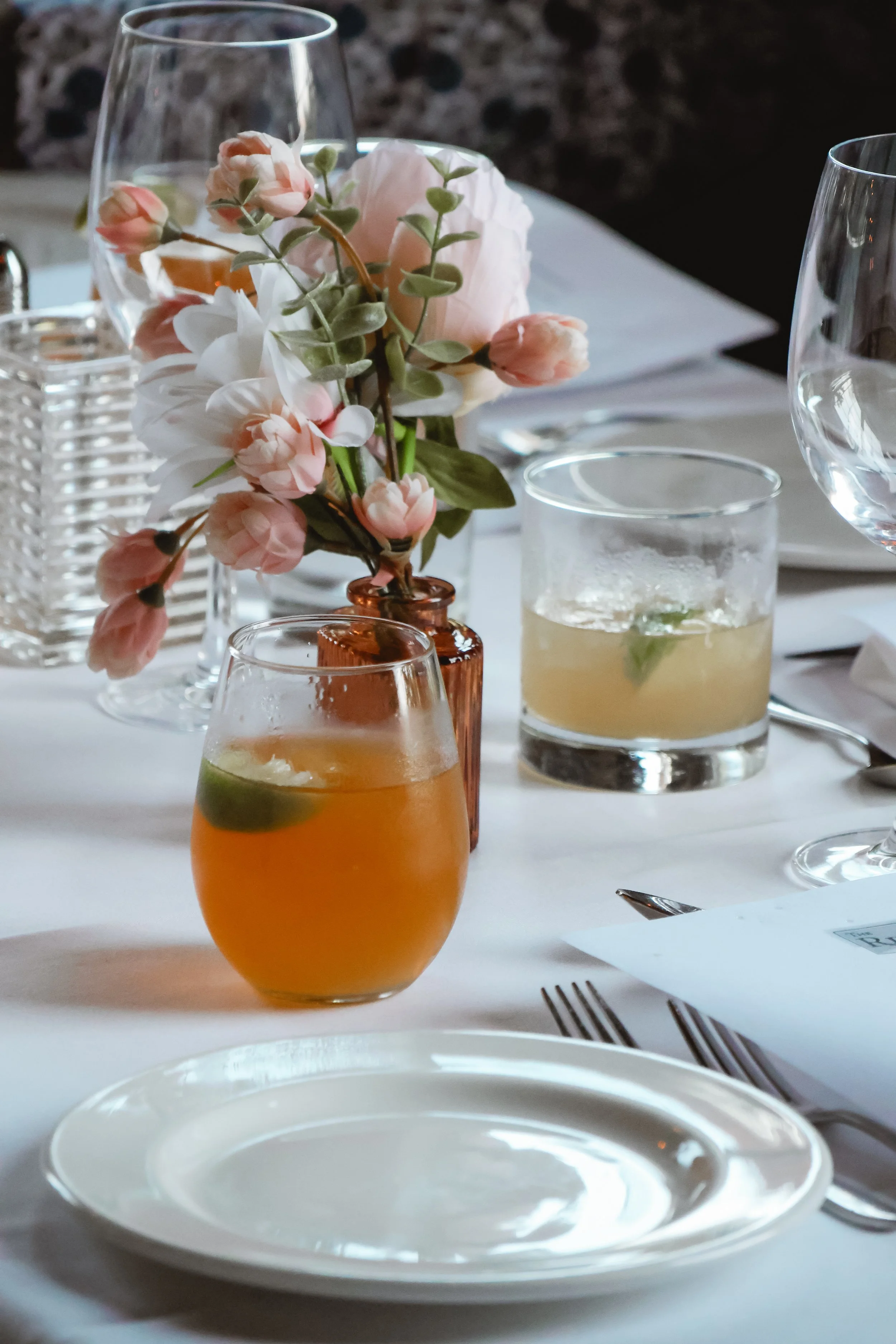 A table setting with a centerpiece of pink and white flowers in a small amber vase, empty white plates, glasses with drinks, and silverware.