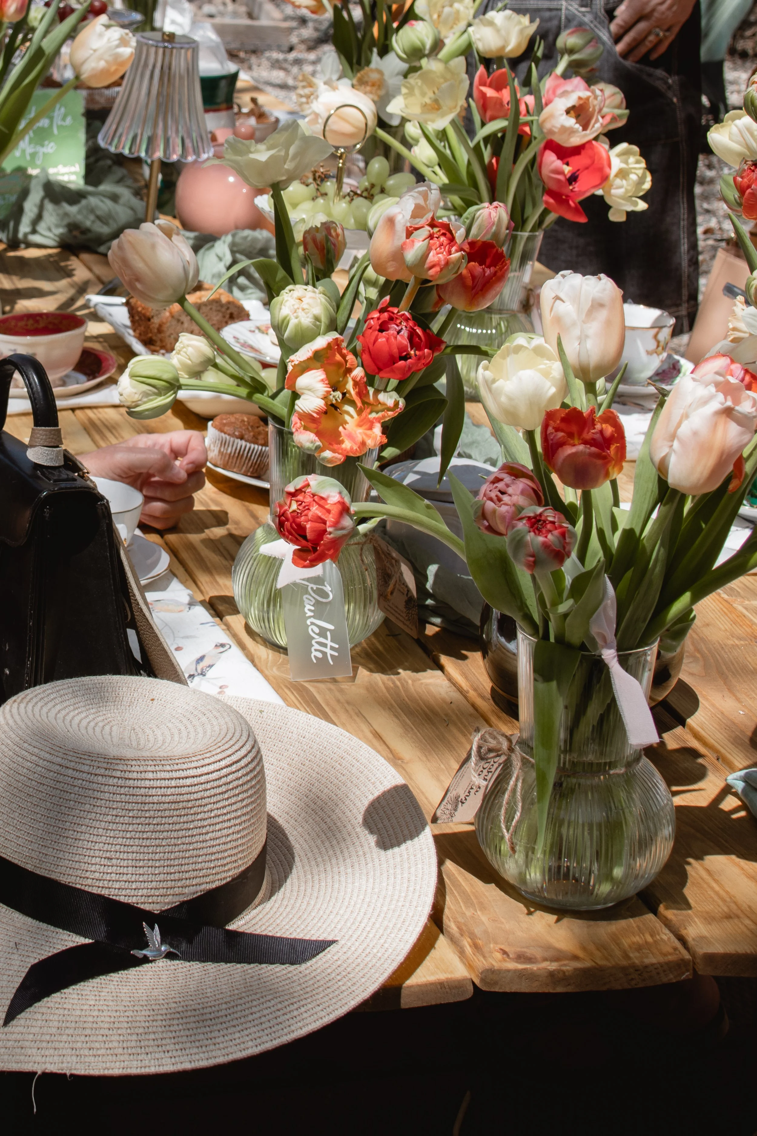 A table decorated with vibrant tulip flower arrangements in glass vases, an elegant straw hat with a black ribbon, and various tableware and food items, suggesting a spring or summer outdoor gathering.
