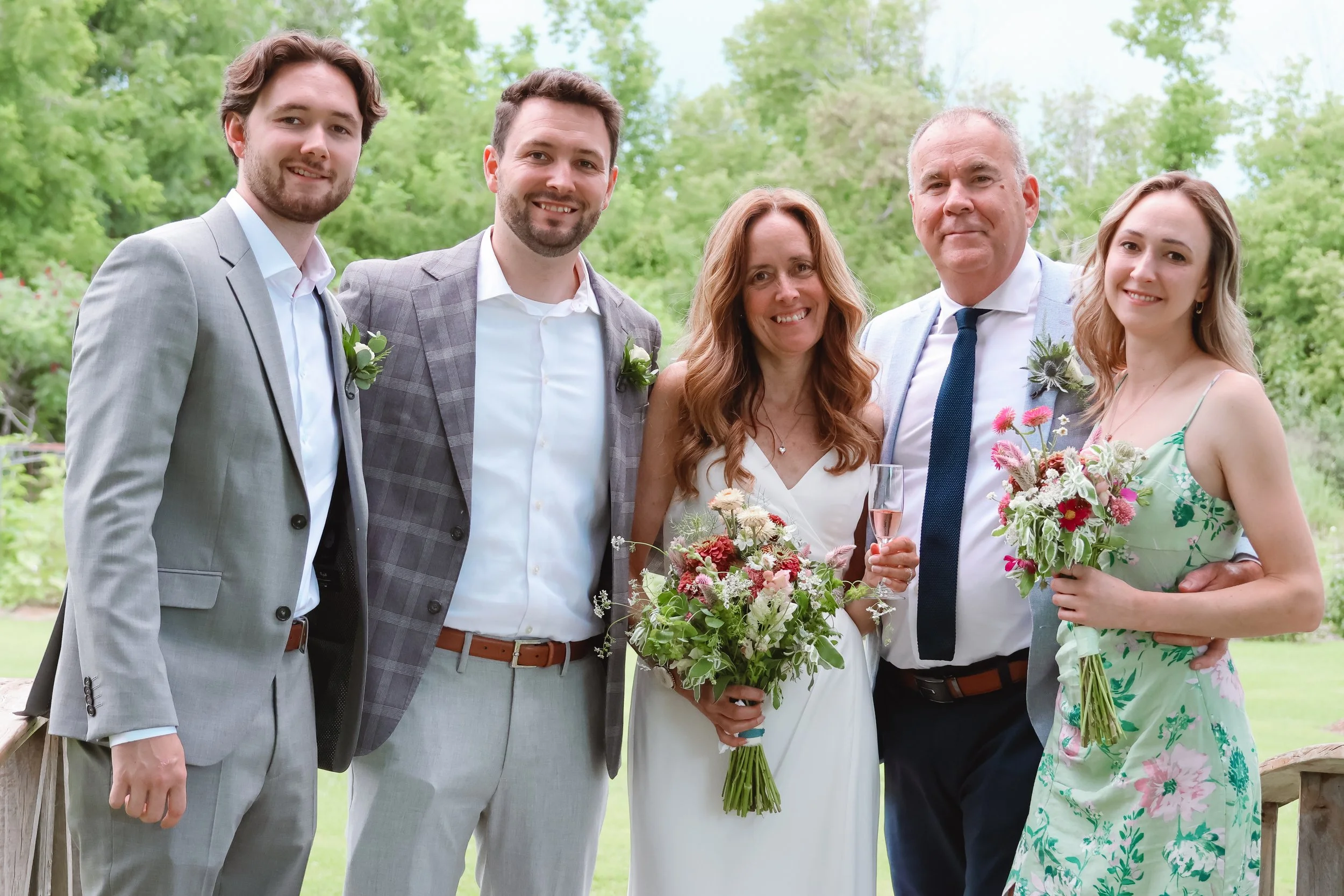 A group of six people, dressed in formal and semi-formal attire, standing outdoors with greenery in the background, celebrating a wedding or special occasion; two women and four men, some holding flowers or champagne.