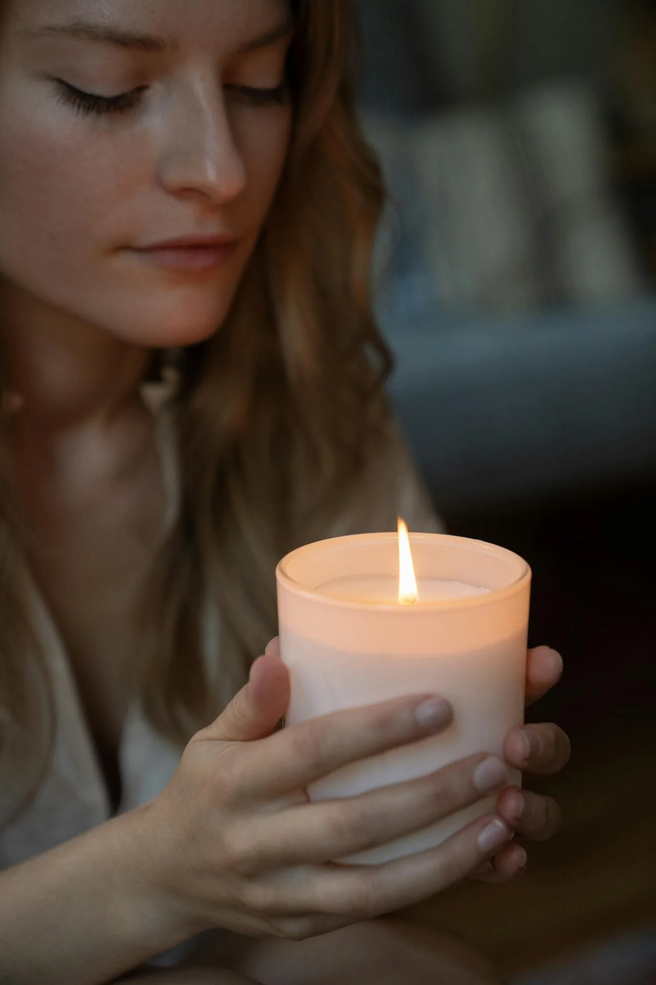 A young woman with closed eyes holding a lit white candle in her hands.
