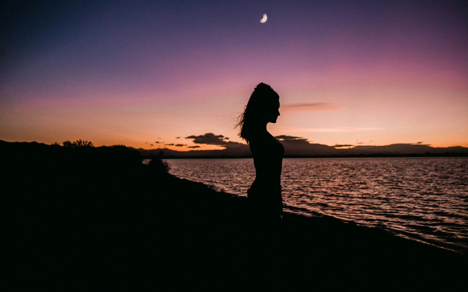 Silhouette of a woman standing by the water's edge during sunset, with a crescent moon in the sky.