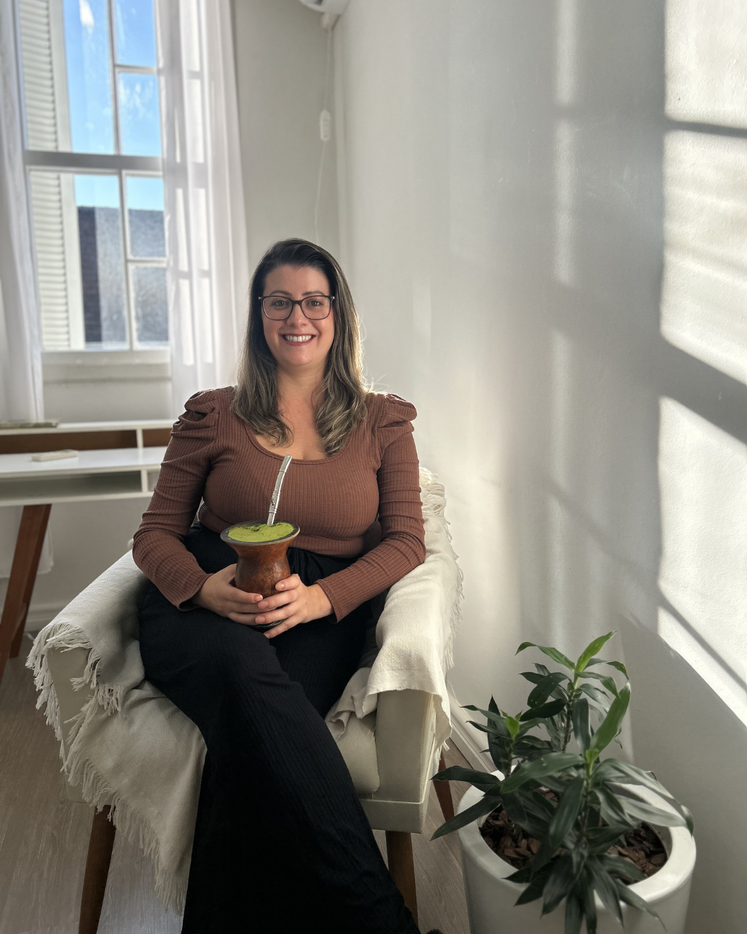 A woman with glasses and long brown hair is sitting in a white armchair in a bright room, holding a traditional mate cup with a metal straw. She is smiling and wearing a brown top and black pants, with sunlight streaming through the window and a potted plant nearby.