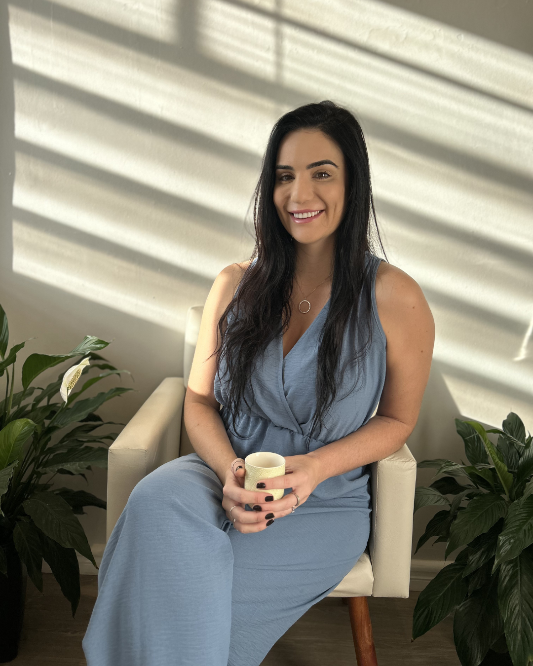 A woman with long black hair, wearing a light blue dress, sitting on a white armchair, holding a mug, smiling, with sunlight and shadows on the wall behind her, and green plants on either side.