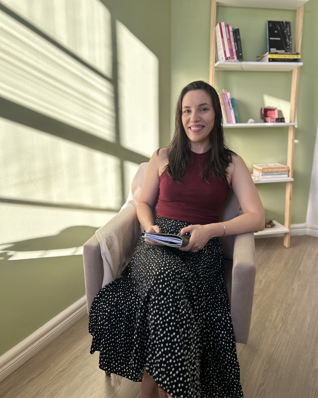 A woman sitting in a beige armchair near a green wall with a bookshelf behind her, holding a notebook, smiling at the camera, with sunlight casting shadows on the wall.