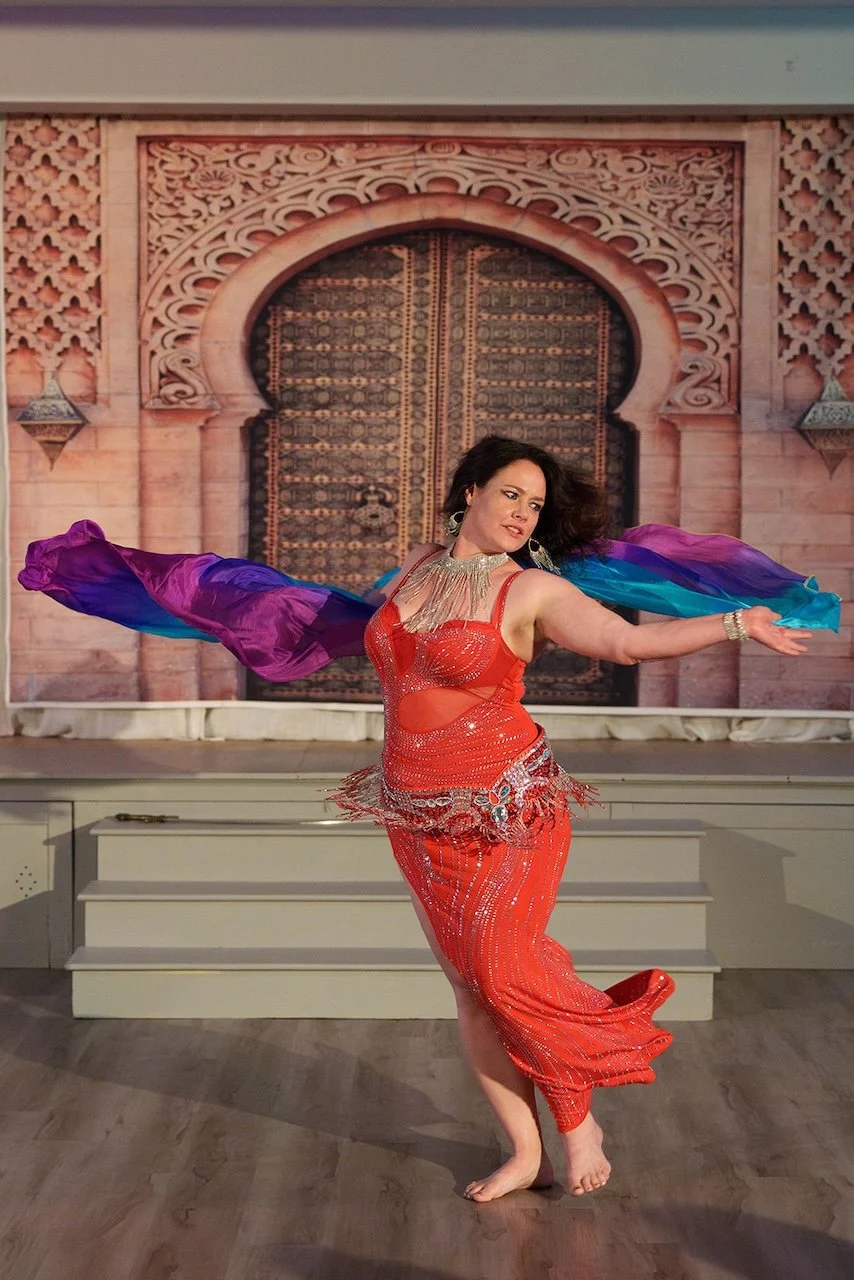 A woman in a red belly dance costume performing on stage with a colorful veil in front of an ornate, decorative backdrop resembling a palace door.