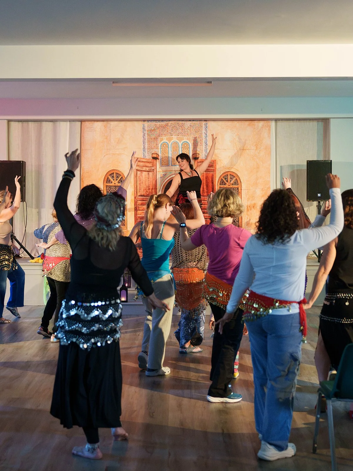 Diverse group of people dancing with belly dancing accessories in a room with a stage backdrop depicting a Moroccan or Middle Eastern scene.