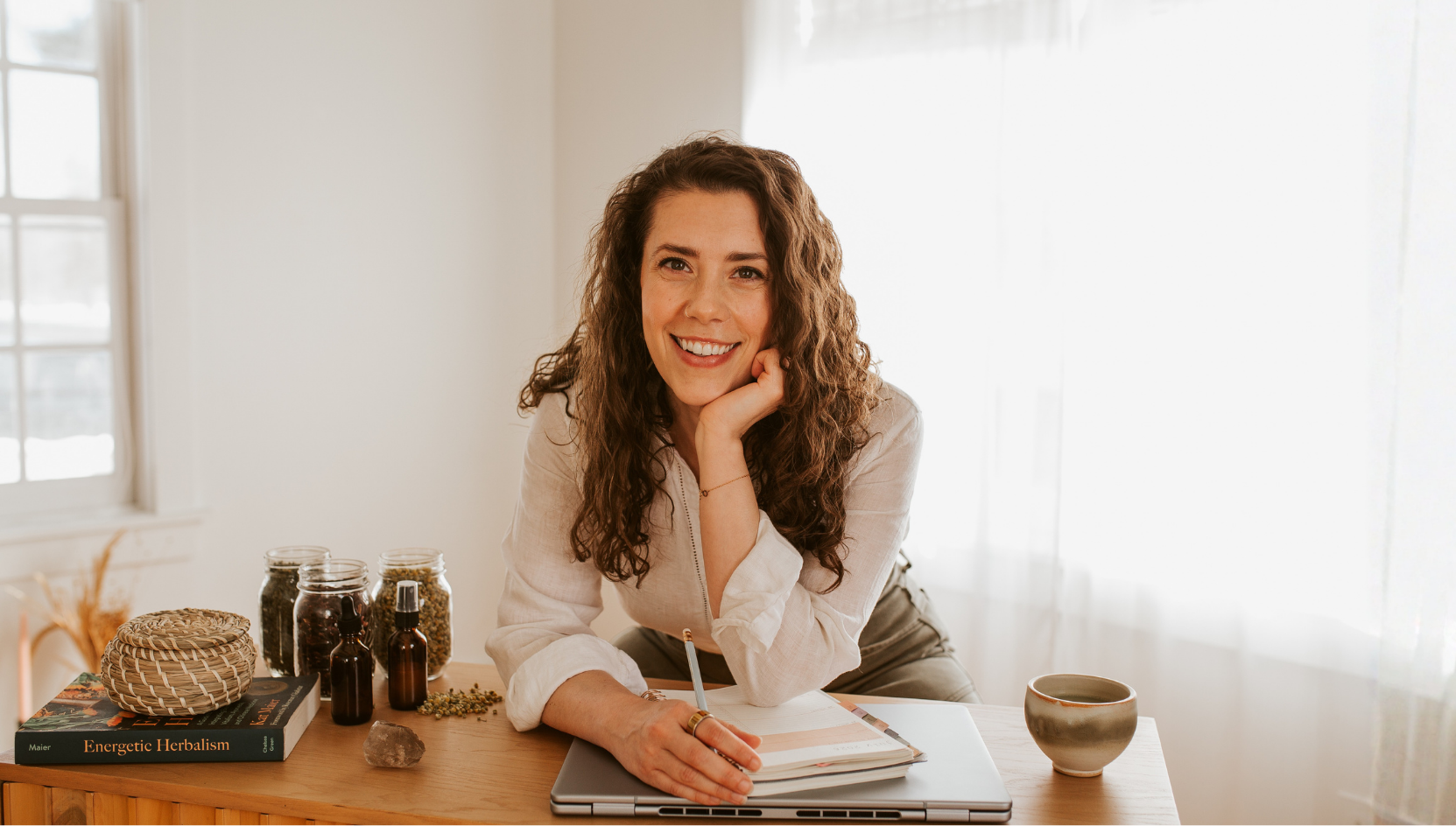 Emily LeDuc, Marketing Strategist, Woman with curly hair smiling at camera while sitting at desk with herbal remedies, a book, a journal, a laptop, and a mug in a bright room.