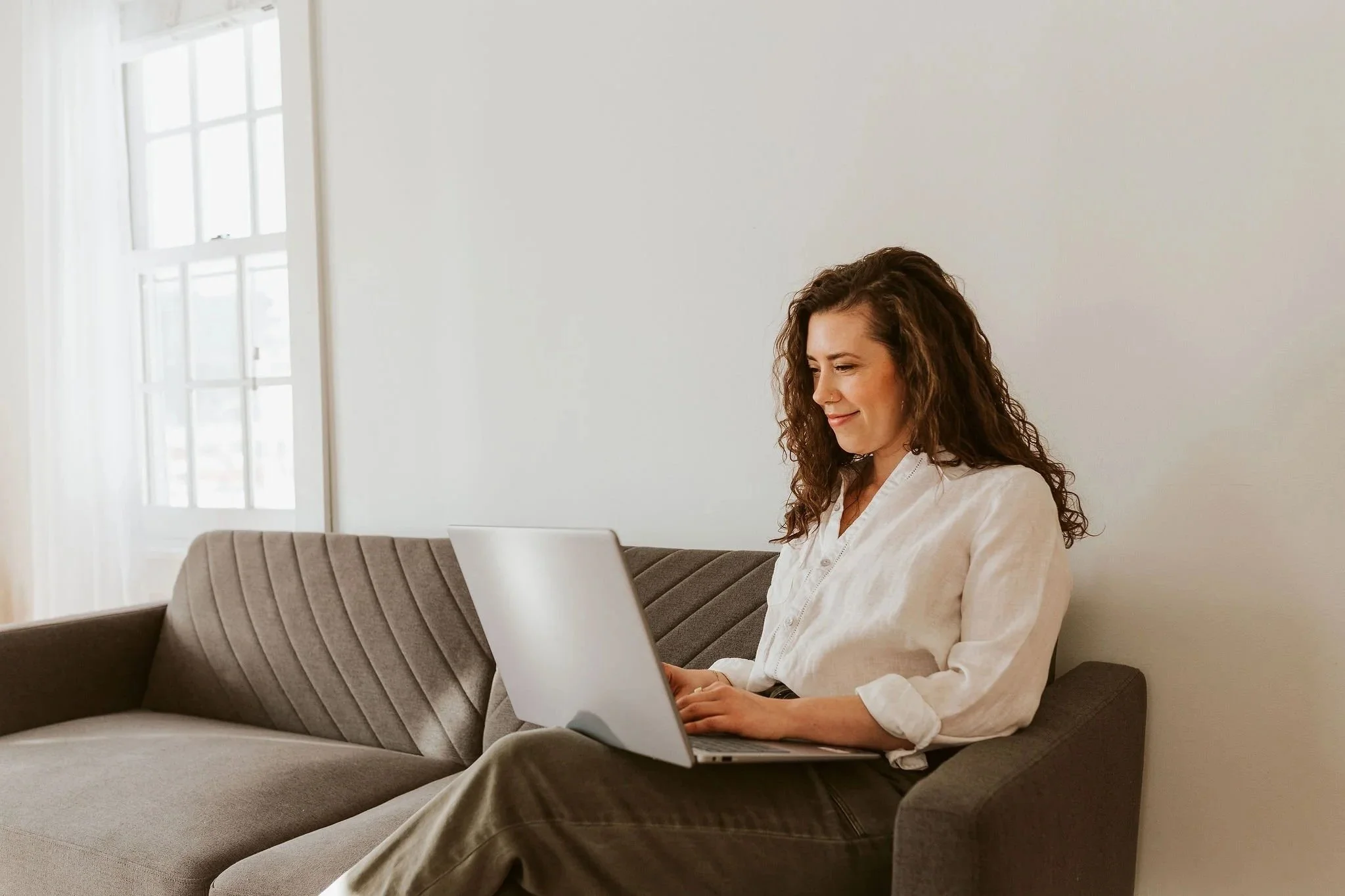 Emily LeDuc, Marketing Strategist, with curly brown hair sitting on a gray sofa, working on a silver laptop, with a white wall and window with curtains in the background.