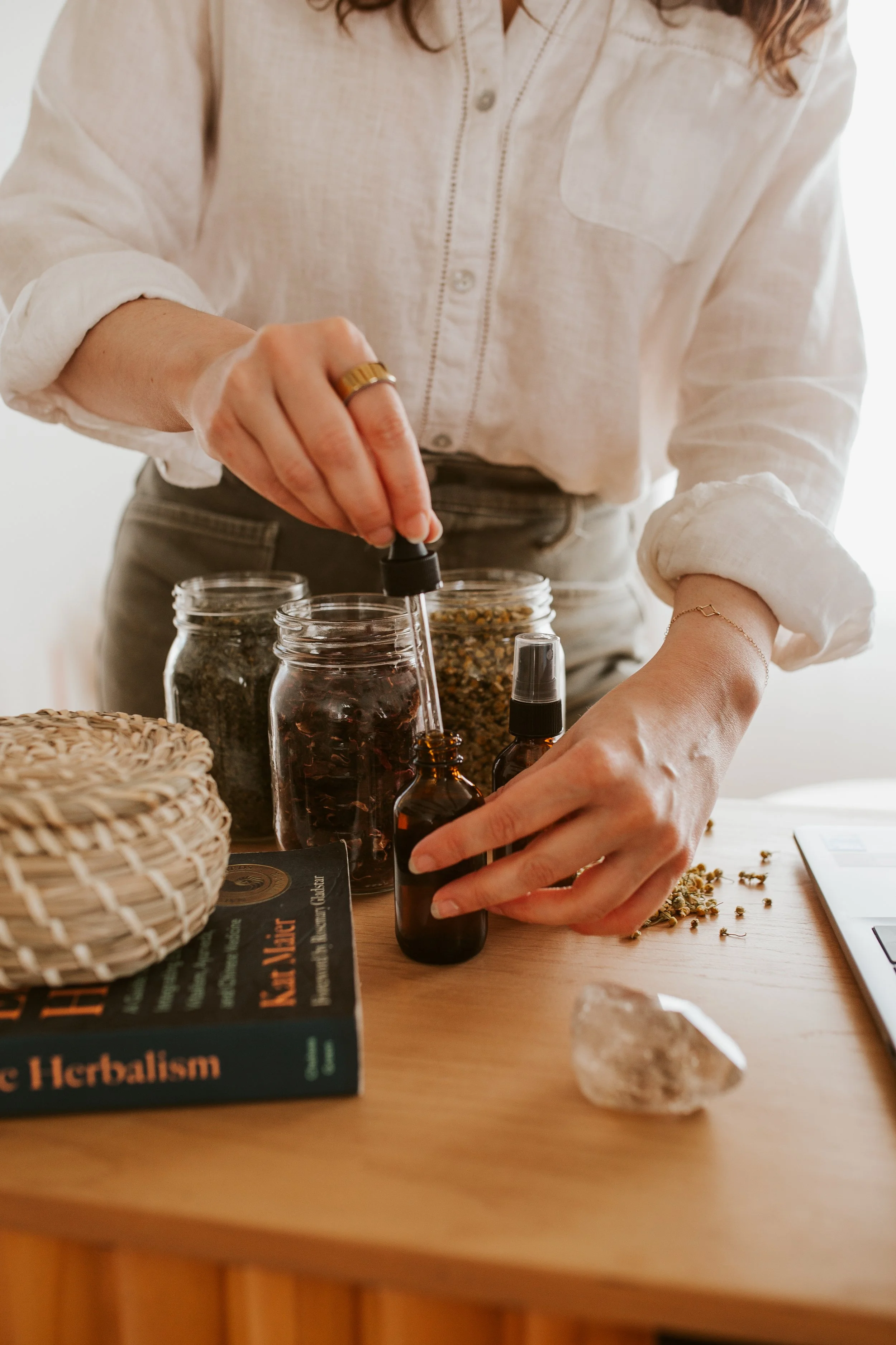 Emily LeDuc -a  person wearing a beige shirt and gray pants is using a dropper to transfer liquid into a small brown bottle on a wooden table. The table also has jars of herbs, a book titled 'Herbalism', a woven basket, scattered dried herbs.