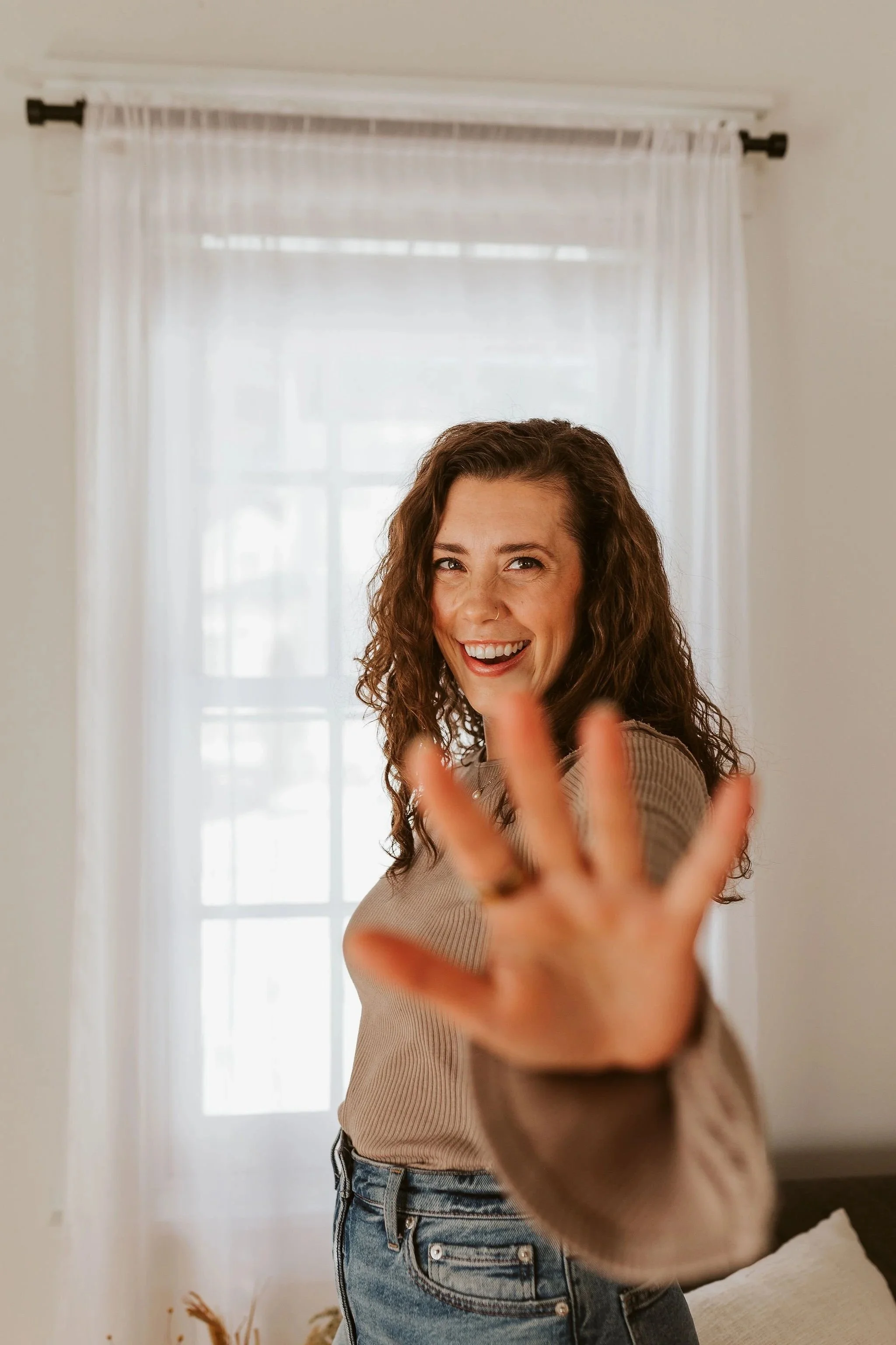 Young woman with curly hair smiling and waving at the camera, standing indoors with a light-colored curtain and window behind her.