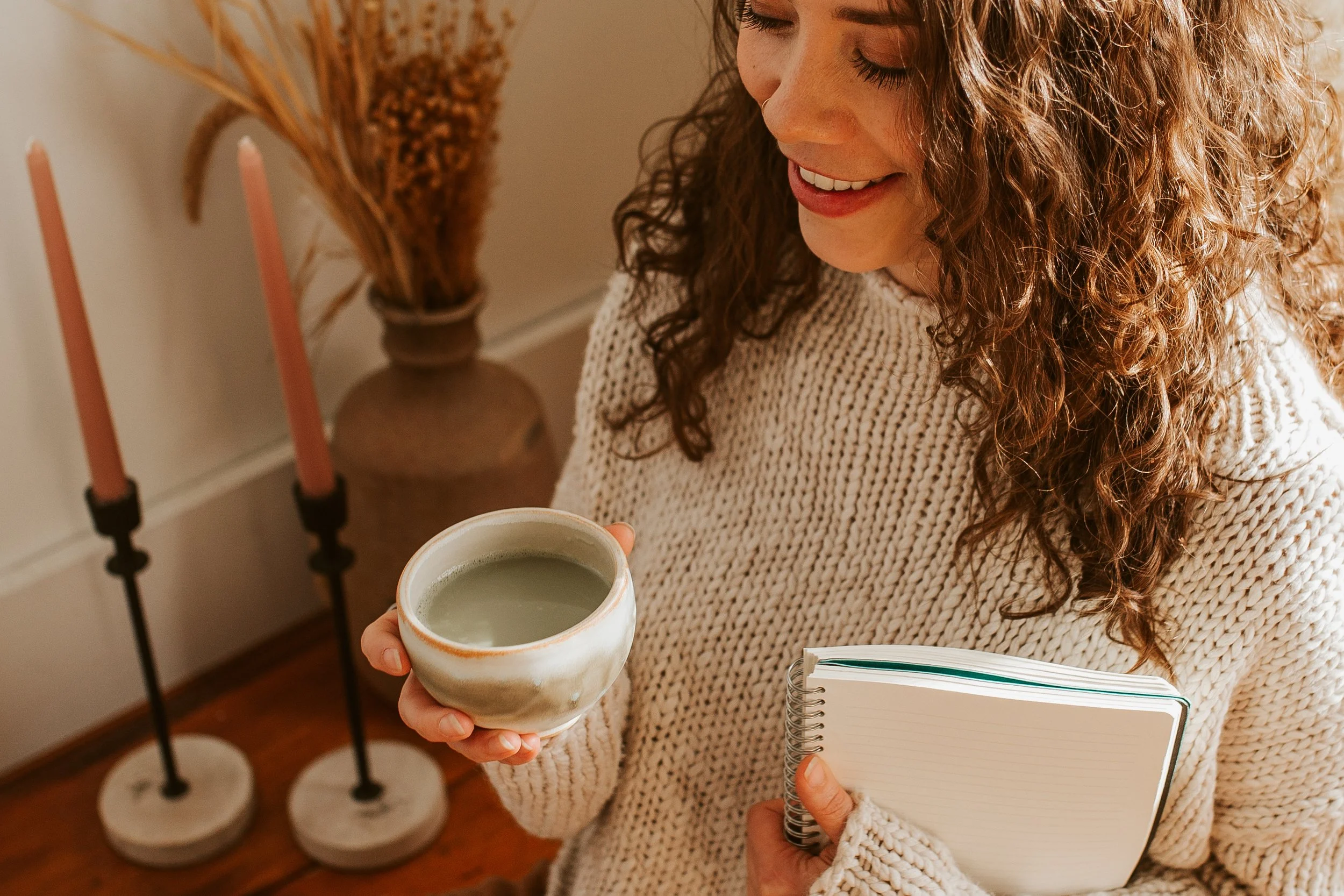 A woman with curly hair wearing a beige knitted sweater, smiling and holding a ceramic cup of green tea in one hand and a lined notebook in the other, standing in a cozy room with a vase of dried flowers and two tall pink candles on black stands in the background.